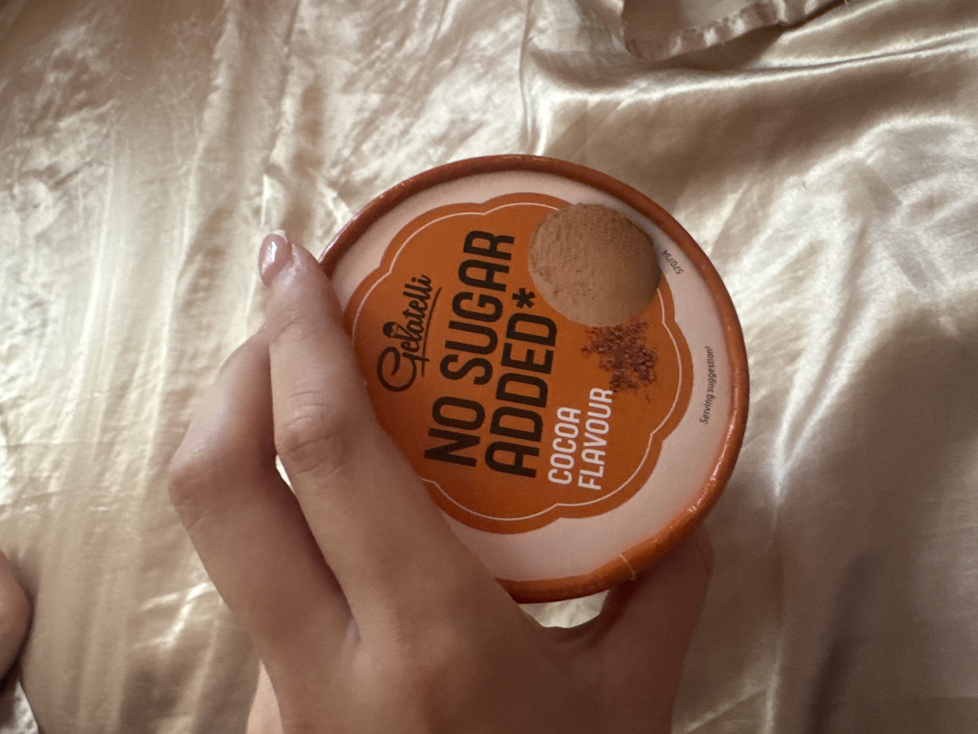The image shows a close-up perspective of a person's left hand holding a round, orange and white container, likely a tub of ice cream or dessert. The hand, with neatly manicured nails adorned with a light, glossy polish, is positioned in the lower left portion of the frame.
The container's lid is prominently featured, displaying the brand name "Gelatelli" in a decorative script. A large, bold declaration "NO SUGAR ADDED*" is central to the design, highlighted in orange on a white background. Below this, the text "COCOA FLAVOUR" specifies the product's taste. The lid also illustrates the product with a realistic depiction of a scoop of cocoa-colored ice cream next to some scattered cocoa powder or chocolate flakes. Small text reading "Serving suggestion" is visible at the bottom right, and a product code, "ST0734," is partially visible on the right edge of the lid.
The background consists of a soft, light-colored fabric, possibly satin or silk, which is gently rumpled, suggesting an indoor setting like a bed or a comfortable seating area. The lighting is even and soft, indicating an interior environment. The image captures a casual moment of someone preparing to enjoy a dessert, potentially within a home in Plovdiv, Bulgaria.