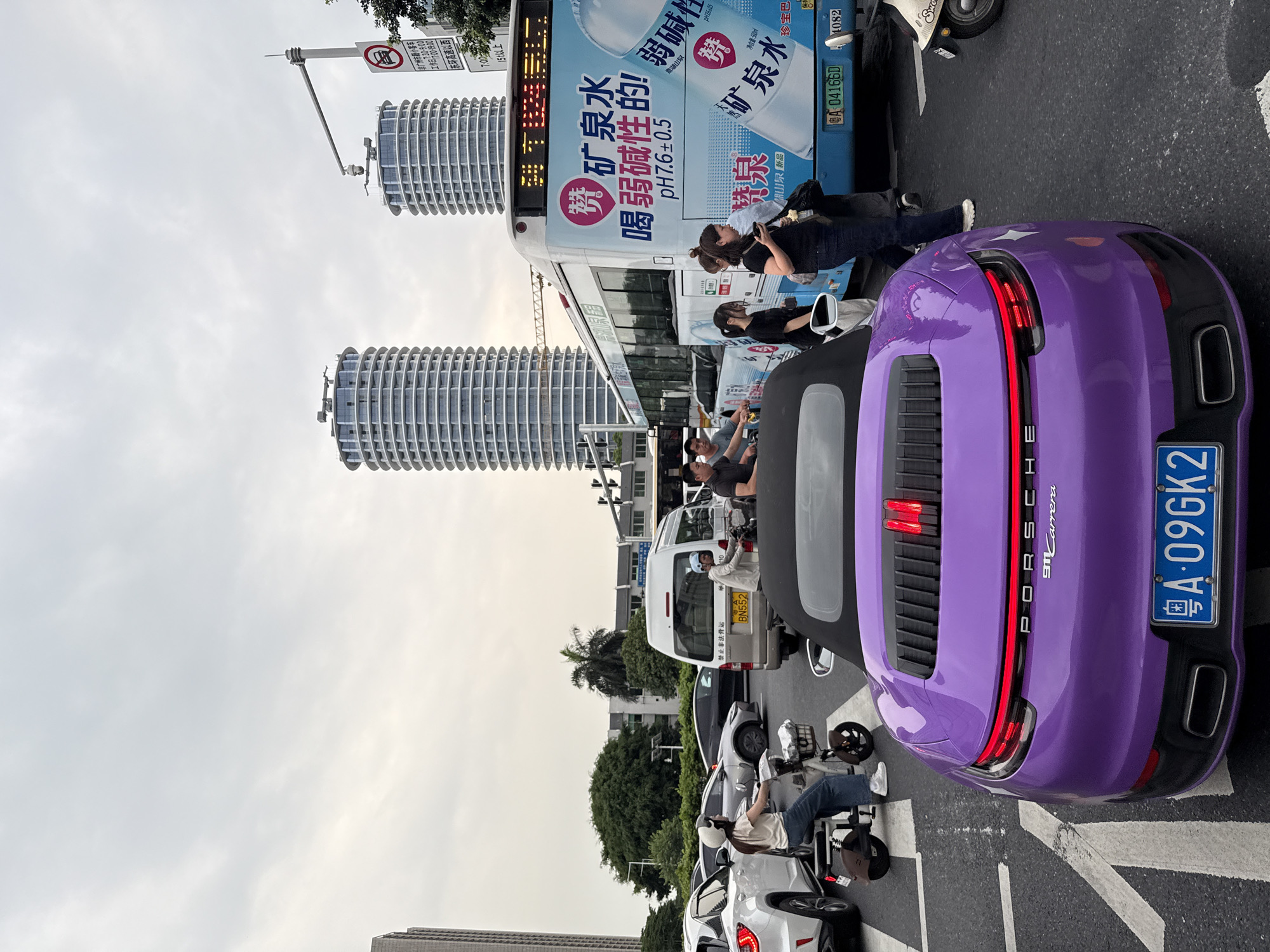 The image depicts a street scene in Tianhe District, China, characterized by a traffic intersection under an overcast sky. In the foreground, a purple Porsche 911 Carrera is parked facing the viewer, its taillights illuminated. To its left, a white SUV is visible. Behind the Porsche, a bus with advertisements for "矿泉水" (mineral water) is stopped, with several people boarding or disembarking. Other vehicles, including a silver van and several motorbikes, are also present in the traffic. Two distinctive cylindrical buildings, appearing to be modern high-rises, dominate the background. A traffic light is visible, as is a street sign with Chinese characters and a red circular symbol with a diagonal line through a vehicle, indicating a restriction. The overall environment suggests a busy urban setting.