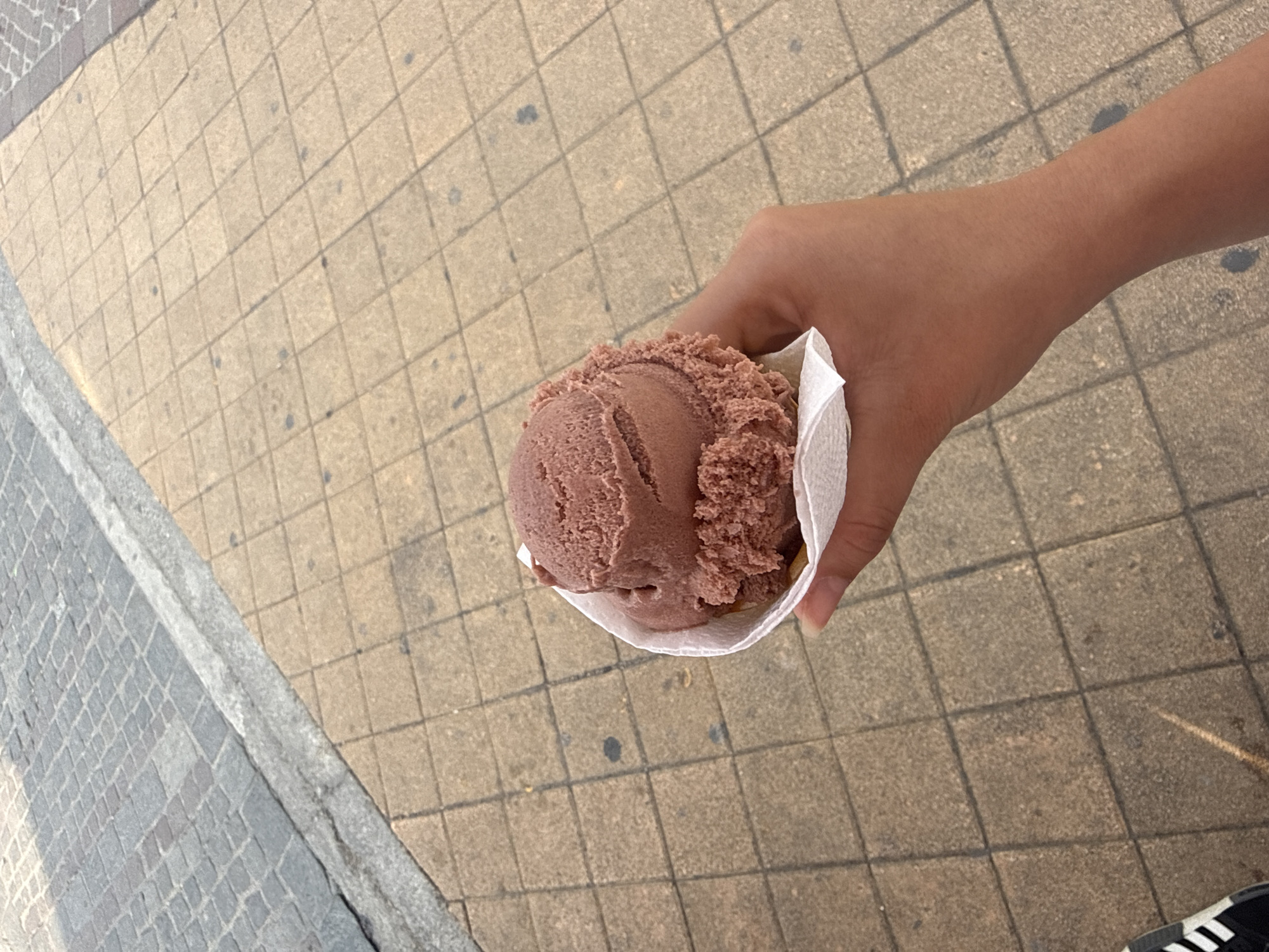 An overhead shot shows a person holding a scoop of chocolate ice cream in a napkin, likely on a city street in San Andrés Cholula, Mexico. The person's hand is visible, holding the ice cream. A portion of a shoe with white stripes is visible in the bottom right corner of the frame. The background consists of a paved sidewalk with square tiles. A curb edge is visible in the left side of the frame.
