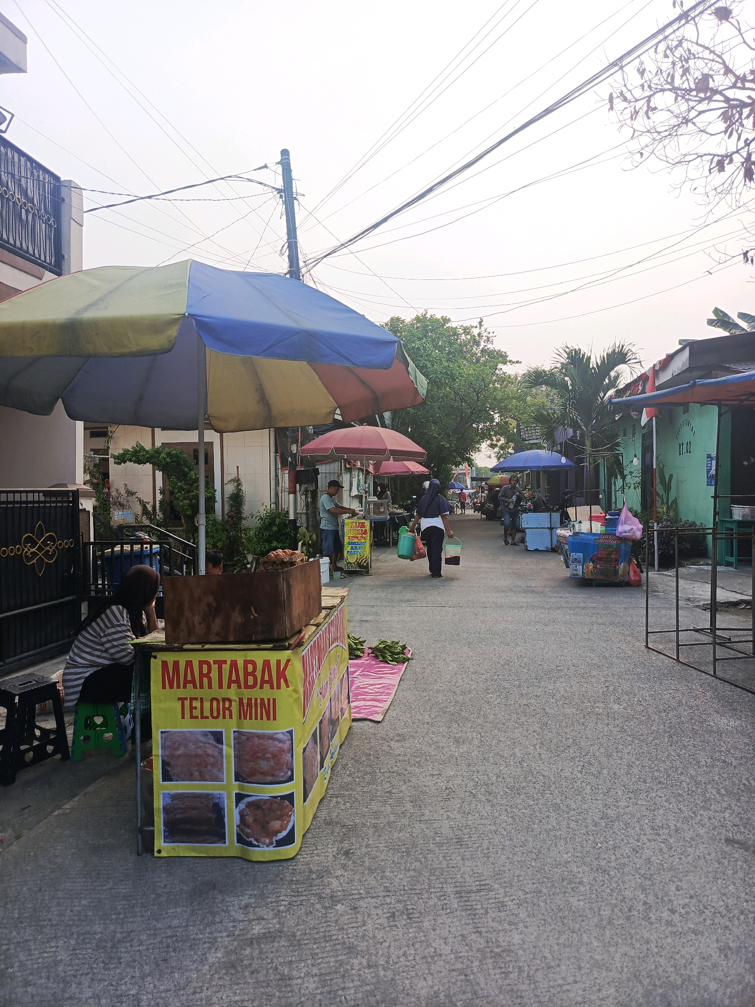 The image depicts a street scene in Balaraja, Indonesia, likely during daylight hours given the illumination. The foreground is dominated by a food stall advertising "MARTABAK TELOR MINI" with images of the dish displayed on a yellow banner. A woman wearing a hijab and a striped shirt sits near the stall, appearing to be observing. To the left, a metal gate with a decorative pattern is visible.
Further down the street, various other stalls and shops line the pavement, some covered by umbrellas of different colors. People are seen moving about, engaged in various activities, including what appears to be a vendor attending to customers at another stall. The street itself is paved with concrete. Overhead, a complex network of electrical wires crisscrosses the sky, supported by poles. Trees and vegetation are interspersed among the buildings, adding a touch of greenery to the urban environment. The overall atmosphere suggests a bustling local market or street food area.