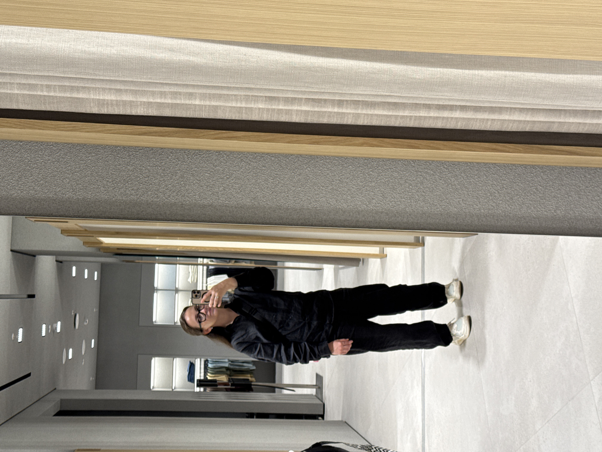 A person is standing in a store taking a selfie in a mirror. The person is wearing glasses, a dark top, dark pants, and white shoes. The store has clothing racks and lighting fixtures. The store's interior is modern with light-colored walls and flooring. The location is unknown, but the user requested Nizhny Novgorod, Russia, which cannot be confirmed from the image.