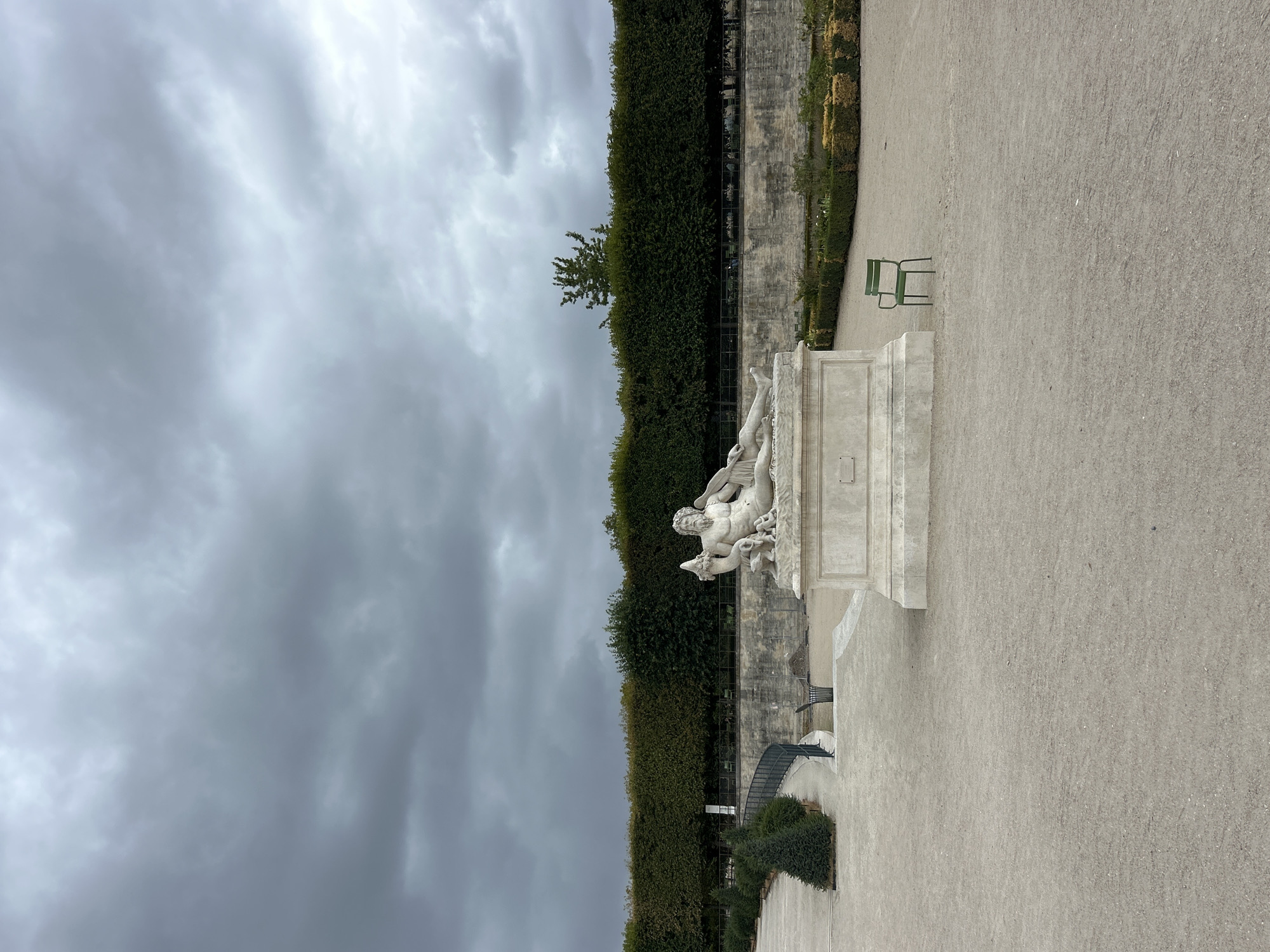 The image depicts a statue of a reclining man on a pedestal in a park. The statue is made of white marble and is adorned with a beard and long hair. The man is depicted in a relaxed pose, with one arm resting on his lap and the other extended upwards. He is accompanied by a winged creature and a dog. In the background, there is a manicured hedge and a stone wall. A single green chair sits in the foreground. The sky is overcast with dark, dramatic clouds, suggesting an impending storm. The scene appears to be the Tuileries Garden in Paris, France, a public garden located between the Louvre Museum and the Place de la Concorde.