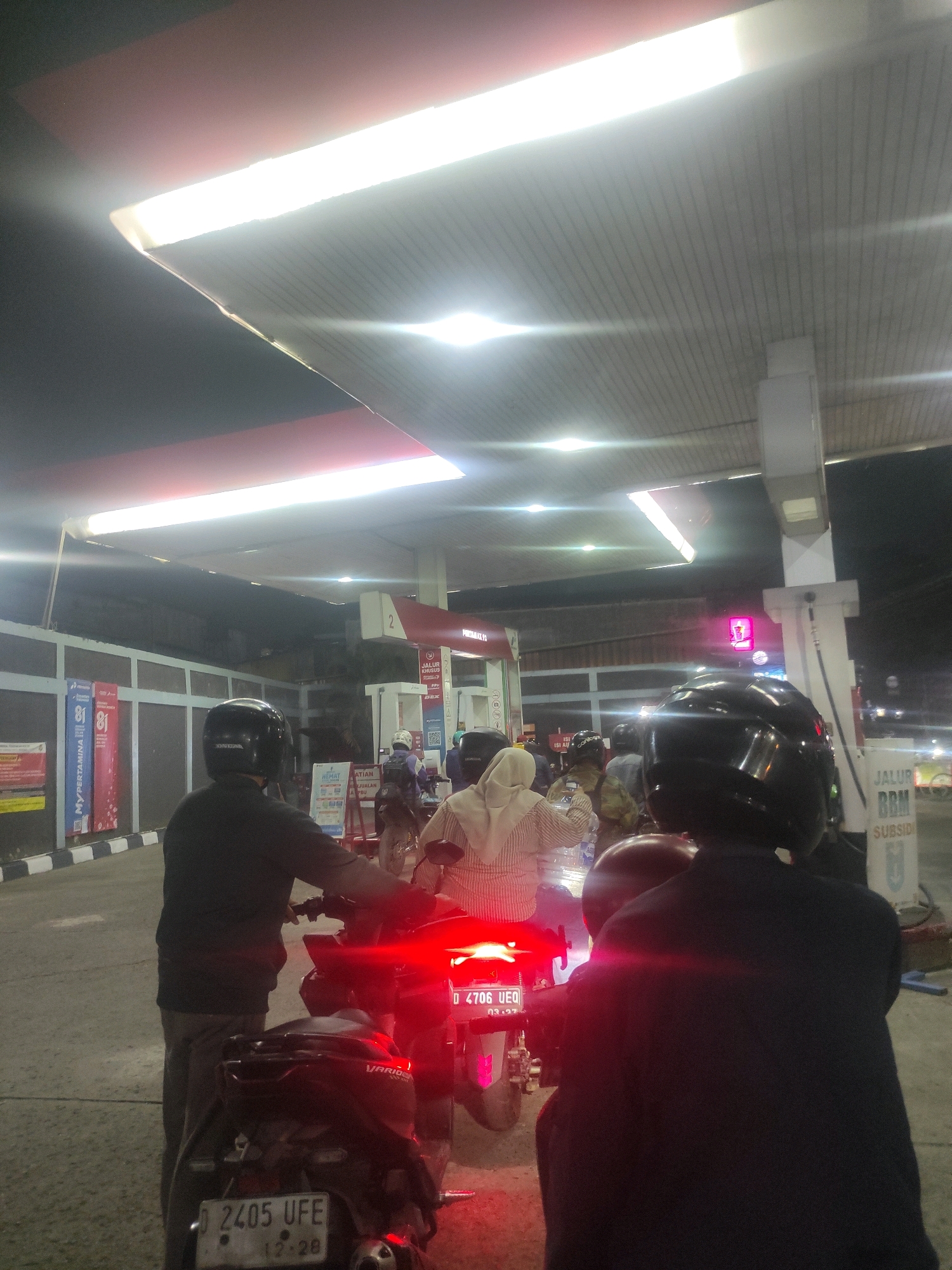 The image depicts a nighttime scene at a Pertamina gas station in Kertamulya, Indonesia. Several people on motorcycles are waiting in line, likely to refuel. The gas station has a large, brightly lit canopy overhead, with fluorescent lights illuminating the area. Red and white signs are visible, including one that reads "JALUR BBM SUBSIDI" (Subsidized Fuel Lane). The motorcycles have license plates, one of which reads "D 2405 UFE" and another "D 4706 UEQ". Some individuals are wearing helmets, and one person is wearing a hijab. The overall atmosphere appears to be a typical evening at a busy gas station.