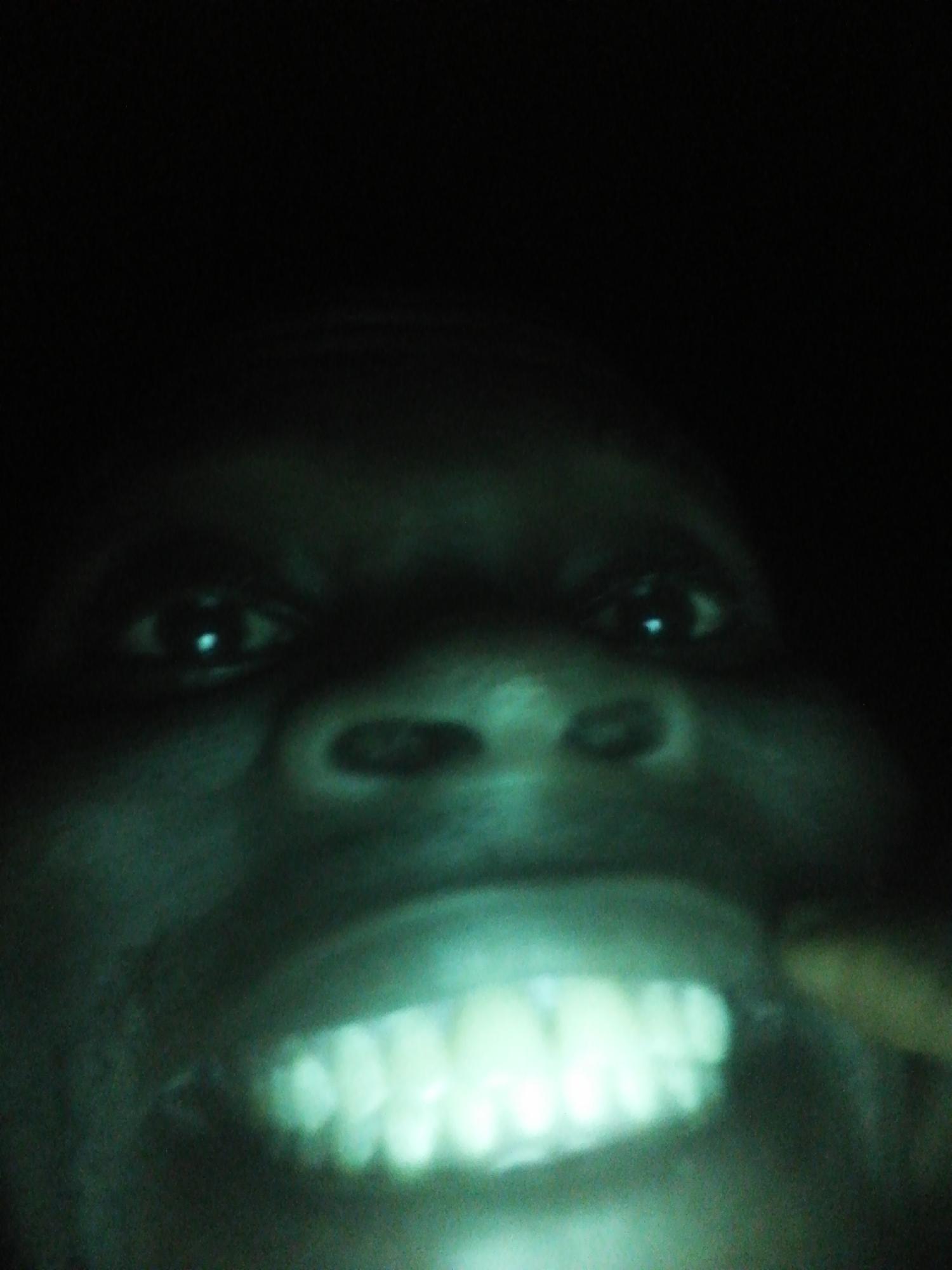 The image shows a close-up of a person's face in low light, with their teeth prominently displayed. The person has dark skin and appears to be looking directly at the camera. The background is dark and indistinct. The location is unknown.