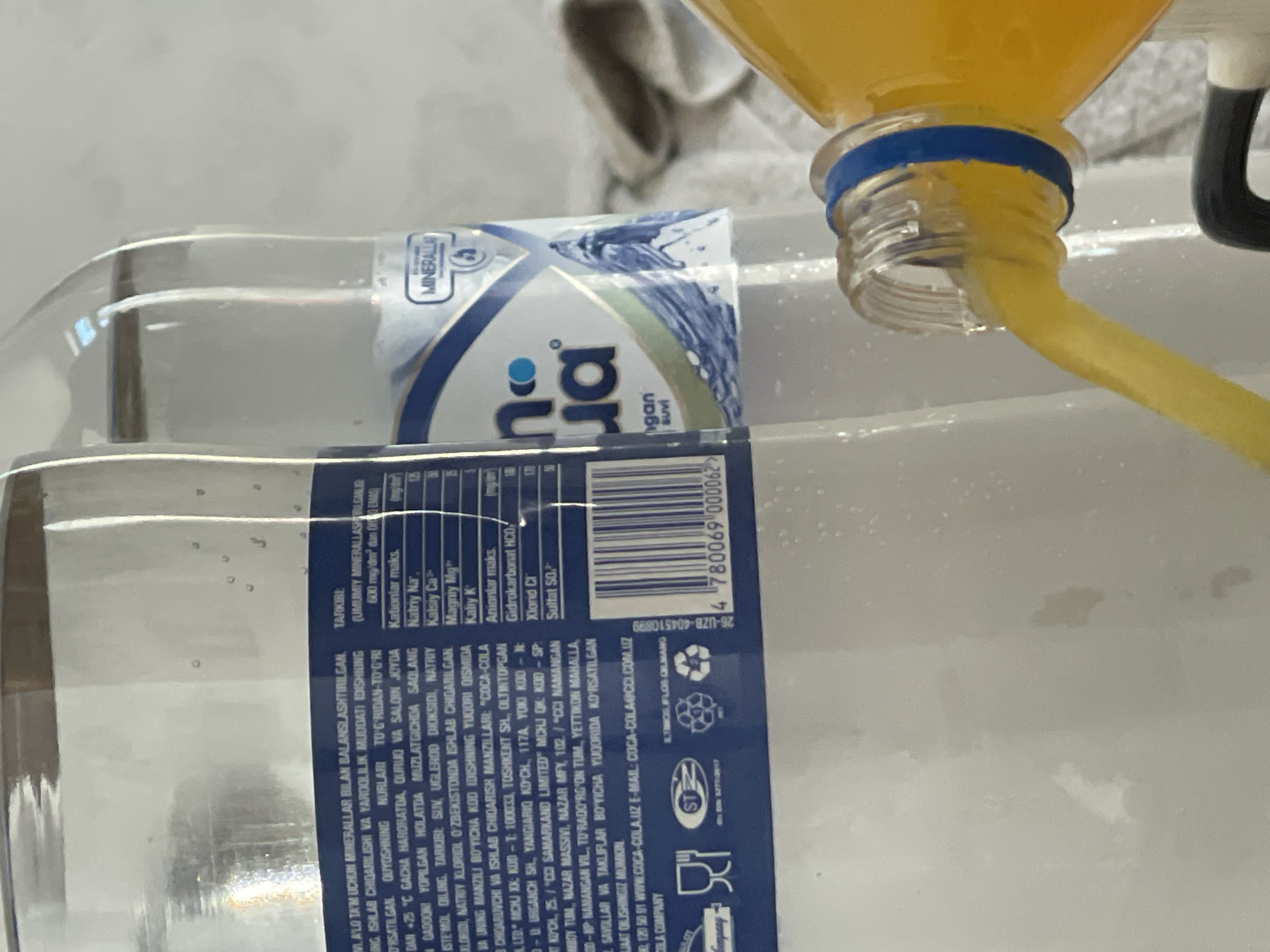 The image shows two plastic bottles of mineral water, one labeled "Bon Aqua" and the other with Cyrillic and Latin script. The "Bon Aqua" bottle has a blue and white label featuring water droplets and a logo that appears to be a wave or waterfall. The other bottle, likely a product of Coca-Cola, has text in a language that appears to be Uzbek. A barcode is visible on this bottle, with the number 4780069000062. The bottles are positioned on a flat surface, possibly a table, with a bright, diffused light source from the left. In the foreground, an orange liquid is being poured from another plastic bottle into an unseen container, with a blue band visible around the neck of the pouring bottle. The background is blurred and appears to be a domestic or commercial setting. There is no visible indication of the city of Tashkent, Uzbekistan, within the image content itself, however, the presence of Uzbek language on one of the bottles suggests a possible location within Uzbekistan.