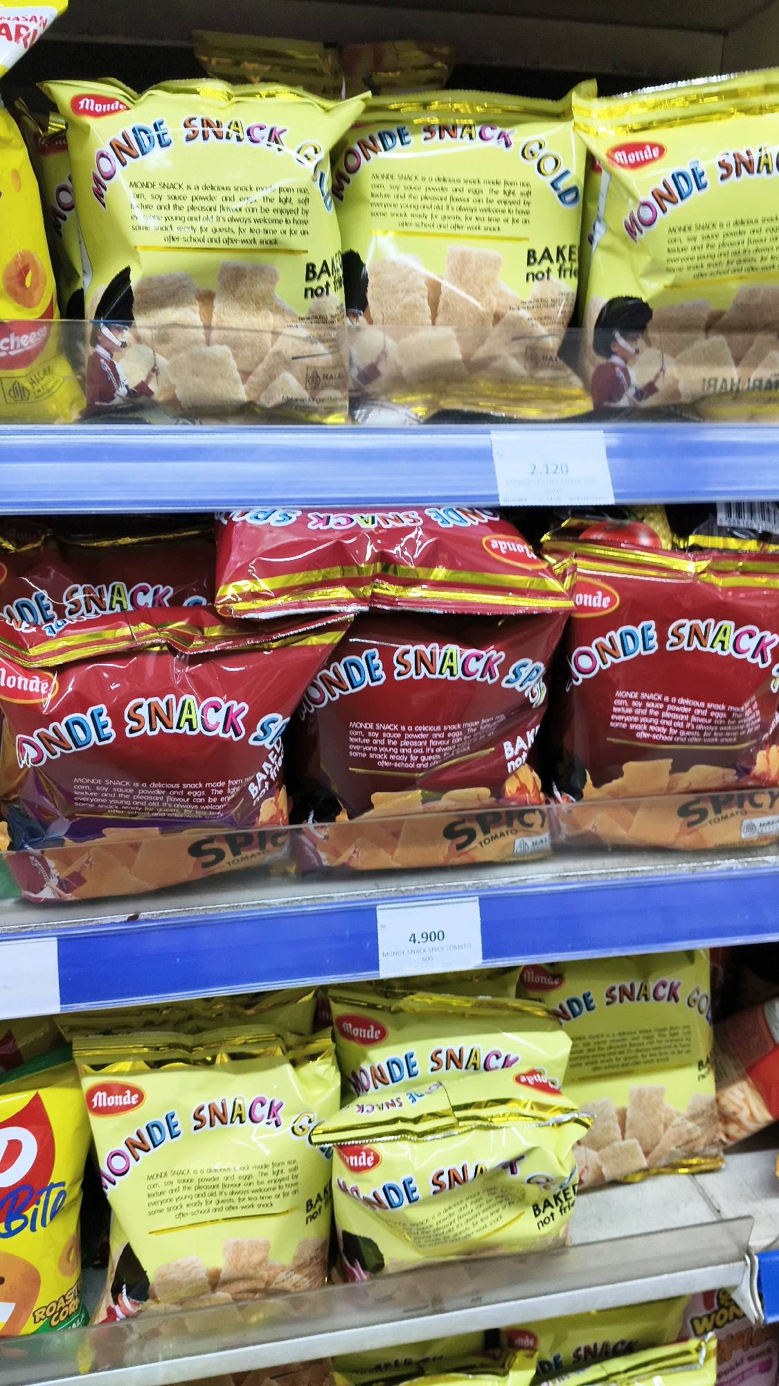 A rack of packaged snacks is displayed on shelves in a supermarket. The snacks are in yellow and red bags with the brand name "MONDE SNACK" prominently displayed in a stylized font. Some bags also feature the words "GOLD" or "SPICY TOMATO." The snacks appear to be small, golden-brown crackers. The shelves are blue and made of metal. The overall environment suggests a retail setting, likely a grocery store or minimarket. Based on the context provided by the user, this scene is located in the city of West Jakarta, Indonesia.
