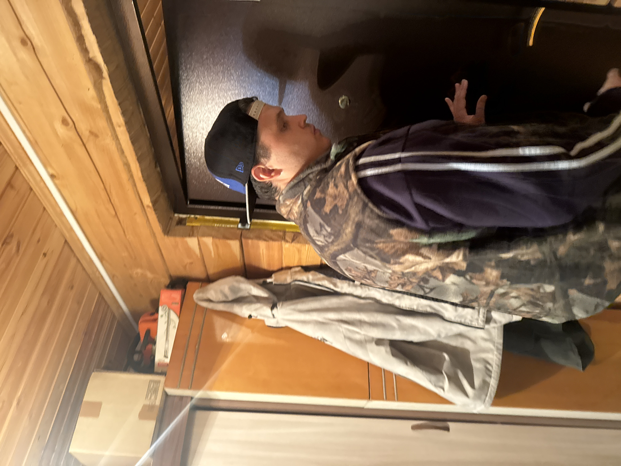 A man wearing a camouflage vest and a baseball cap is standing in what appears to be an indoor space. The space is characterized by wooden paneling on the walls and what looks like a cabinet or drawer unit below the man. There is a light-colored jacket or coat draped over the cabinet. The man's hand is partially visible within a dark area that could be a shelf or storage space above him. A chainsaw in its case is visible on a shelf. The location in Mayak, Russia, is not verifiable from the image.