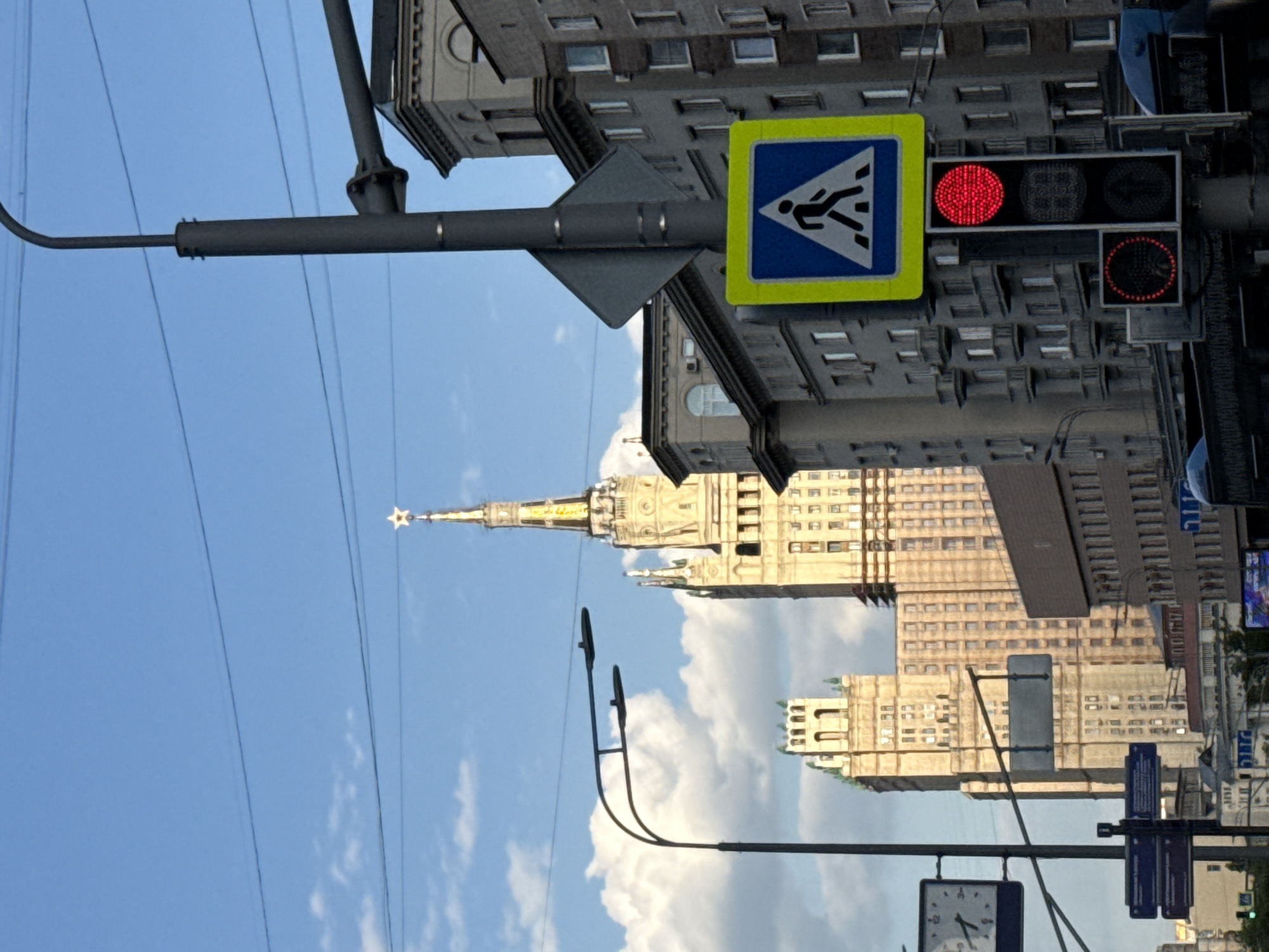 The image presents a daytime street view in Moscow, Russia, showcasing a prominent example of Stalinist architecture under a clear blue sky. A tall, ornate building, characteristic of the city's "Seven Sisters" skyscrapers, stands in the mid-ground, featuring a tiered facade in a light hue, crowned with a spire and a gold star at its apex.
In the foreground, a dark gray utility pole supports several urban fixtures. To the left, a blue pedestrian crossing sign with a white walking figure is clearly visible. Adjacent to it, a traffic light displays a solid red circle, indicating a stop signal for vehicles. Below this, a smaller pedestrian traffic light also shows a red illuminated outline of a walking figure, signaling pedestrians to wait. Overhead wires crisscross the frame against the bright sky.
Other multi-story buildings, likely residential or office blocks, flank the main skyscraper and extend into the background. The sky is a vibrant blue with scattered white clouds, suggesting fair weather and ample sunlight illuminating the scene. The time of day appears to be late morning or afternoon. The activity depicted is a momentary pause in traffic and pedestrian movement, with no specific human figures visible. No discernible text is present on the signs or buildings, only universal symbols.
