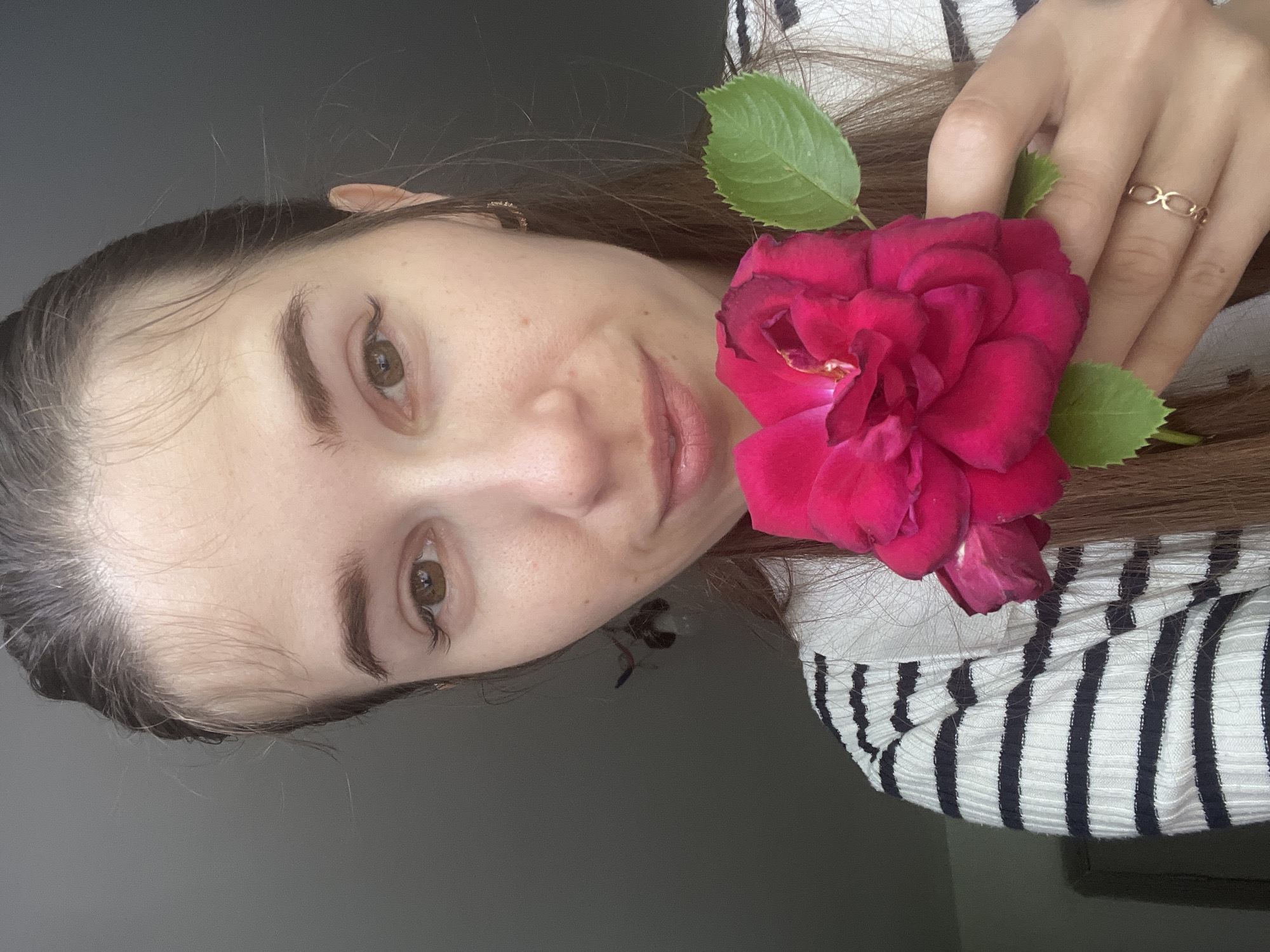 A young woman with brown hair and brown eyes holds a red rose in her right hand. She is wearing a black and white striped sweater. The woman is looking at the camera with a slight smile. The background is a dark, out-of-focus wall.