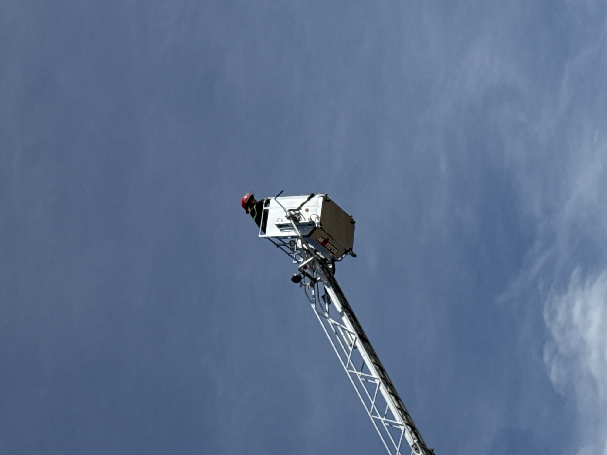 A firefighter, wearing a red helmet and dark clothing, stands in the basket of an elevated fire ladder. The ladder extends upwards diagonally across the frame against a backdrop of a pale blue sky with wispy white clouds. The basket is white and metallic, with some visible equipment and railings. The word "MORITA" is clearly visible in black lettering on the side of the basket. The sky suggests it is daytime, possibly late morning or early afternoon, with good visibility. The scene appears to be an operational scenario or training exercise, with the firefighter positioned high above the ground. There are no discernible location cues specific to Da Lat, Vietnam, beyond the sky which is consistent with any open outdoor setting. The overall mood is professional and focused, consistent with a rescue or emergency response.
