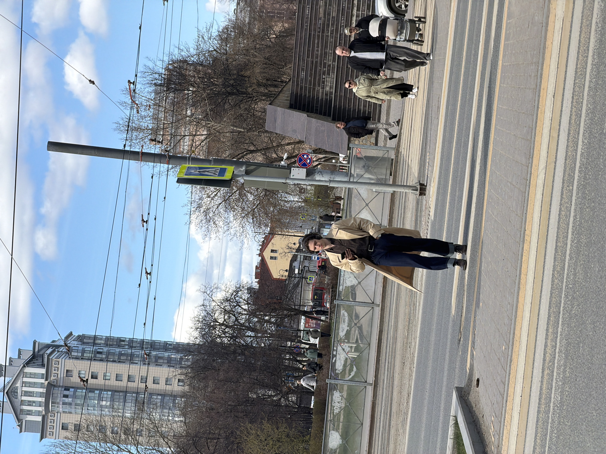 The vertical image, set in an urban environment in Perm, Russia, captures a bright daytime scene under a blue sky with scattered white clouds. The primary subject in the foreground is a person standing on a paved sidewalk, wearing a light-colored trench coat and dark trousers, looking towards the viewer. They are positioned near a pedestrian crossing, indicated by zebra stripes on the asphalt road.
Across the street, several individuals are visible. Three people stand near a set of stone steps and a landscaped area: one in a light beige coat, another in a striped shirt, and a third in a dark jacket. Further back, near a pole and a bus stop-like structure, another person is seen. The street features distinct pedestrian crossing markings and yellow lines separating lanes or areas. A metal fence borders the sidewalk, separating it from a park-like area with numerous bare trees, suggesting either early spring or late autumn.
Prominently featured are utility poles and a network of overhead wires, likely for trams or trolleybuses, crisscrossing the sky. A tall, modern multi-story building with a complex roofline and extensive glass facades dominates the left side of the background. Behind the bare trees, other older, lower-rise buildings with light-colored facades are visible.
Visible signage includes a blue square pedestrian crossing sign (white triangle with a walking person) and a circular 'No Stopping' sign (blue with red border and diagonal line). The overall impression is one of a bustling city street on a clear, cool day.