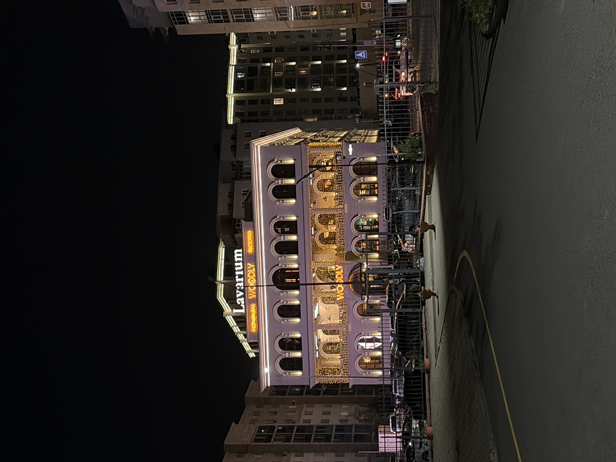 The image shows a night view of a building in Astana, Kazakhstan, identified by the illuminated sign "Lavarium." The building is decorated with numerous string lights, illuminating its facade, which features arched windows and balconies. Below the main building, a sign reads "WOODLY," indicating a restaurant or establishment. In the foreground, a dark asphalt road with a yellow line is visible, leading towards the building. A black metal fence separates the road from a parking area where several cars are parked. Surrounding the central building are other multi-story residential or office buildings, also illuminated. The sky is dark, typical of a nighttime scene.