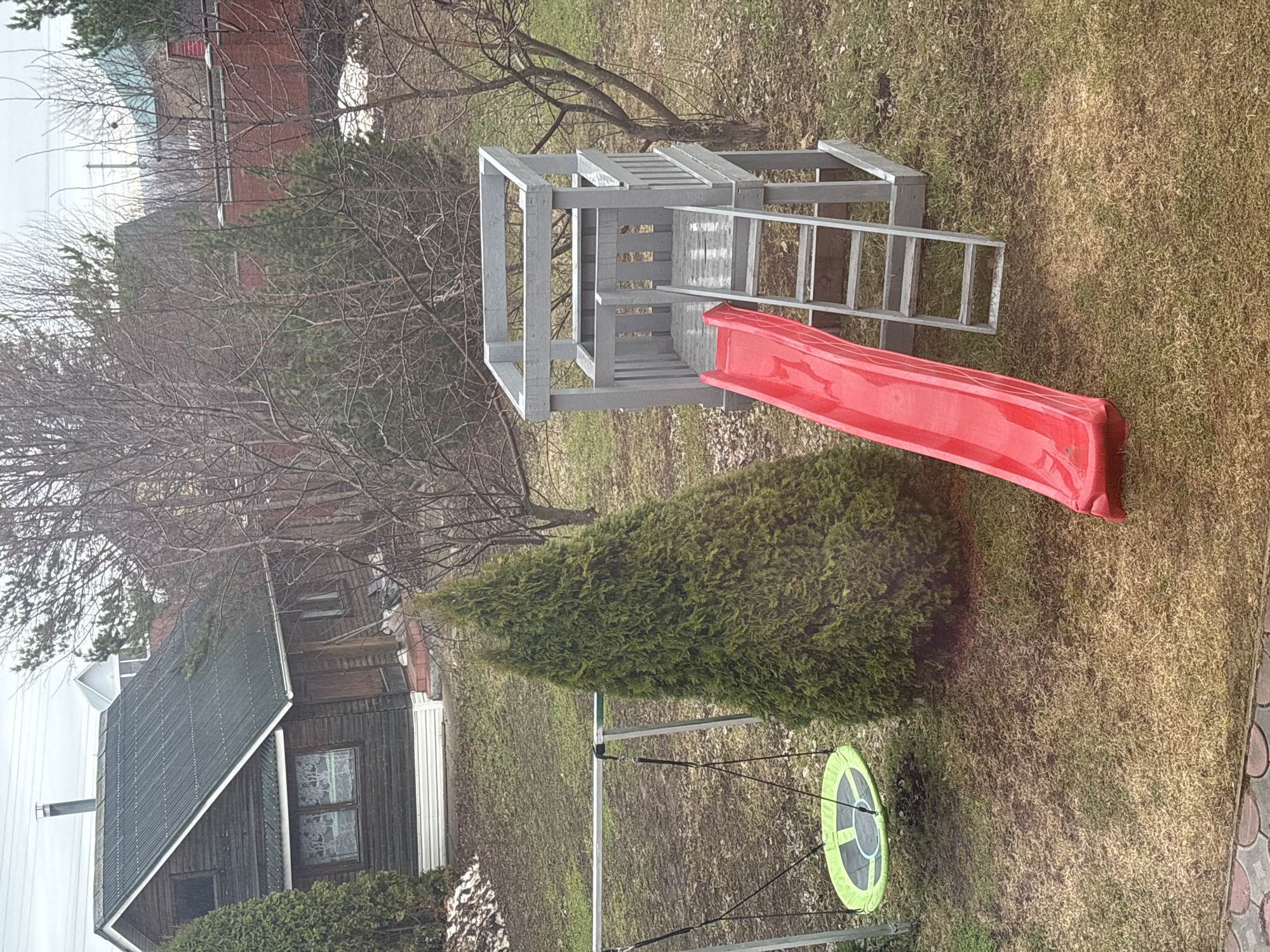 The image depicts a backyard scene in Ферма, Russia. A wooden play structure, painted gray, dominates the scene. The structure includes a ladder, a platform, and a red plastic slide. Near the slide is a conical evergreen shrub. A circular swing, green and black, hangs from a metal frame in the lower right of the frame. The ground is covered in dry, brown grass. In the background, a wooden house with a gray roof is partially visible on the left. Bare trees are interspersed throughout the scene and a brick house can also be seen through the trees in the background. The sky appears overcast.