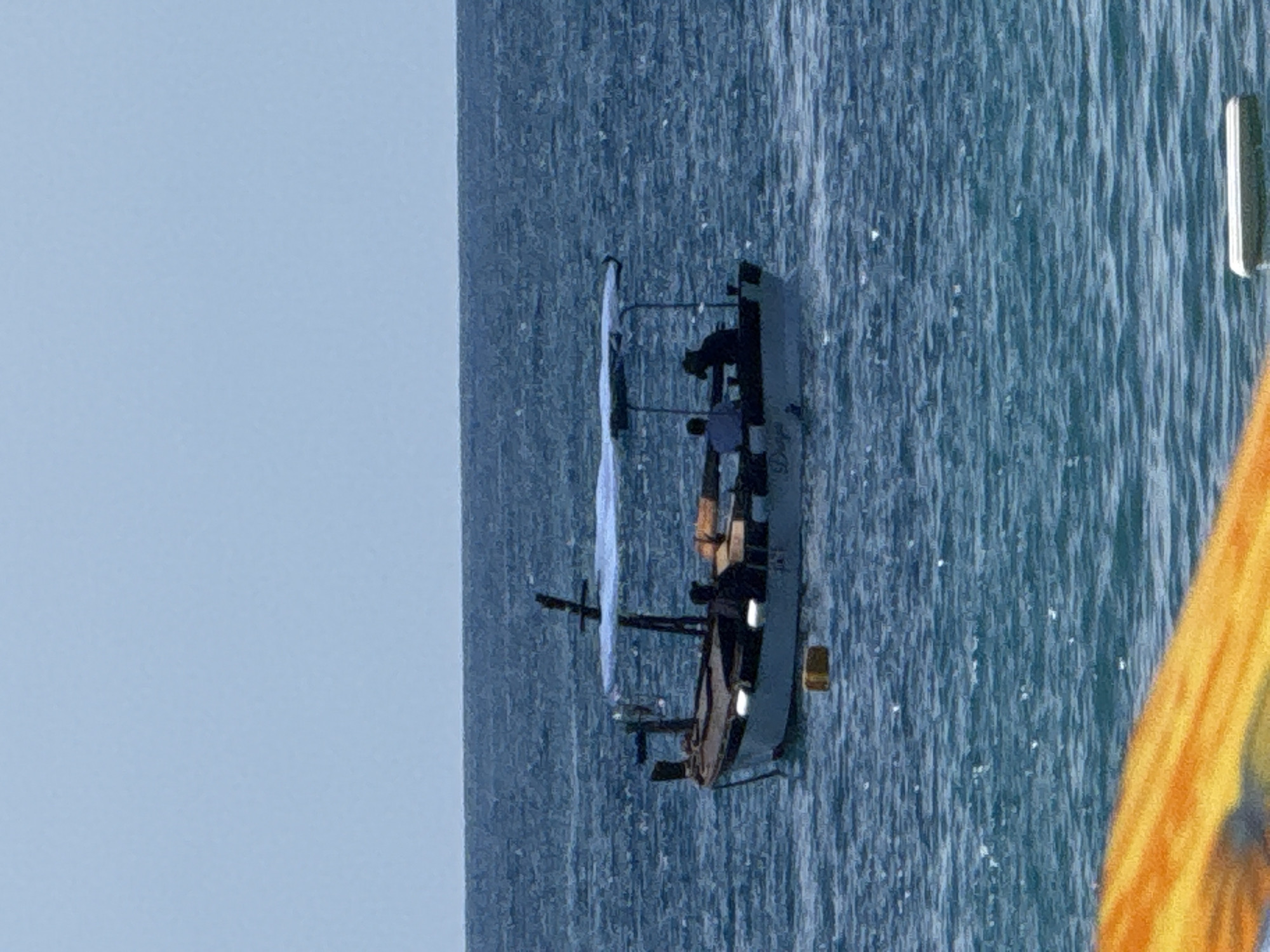 A boat with a blue canopy is on the water. There are at least three people in the boat. One person is wearing a hat. The water is choppy. The sky is clear and blue. In the foreground, a portion of an orange object is visible.