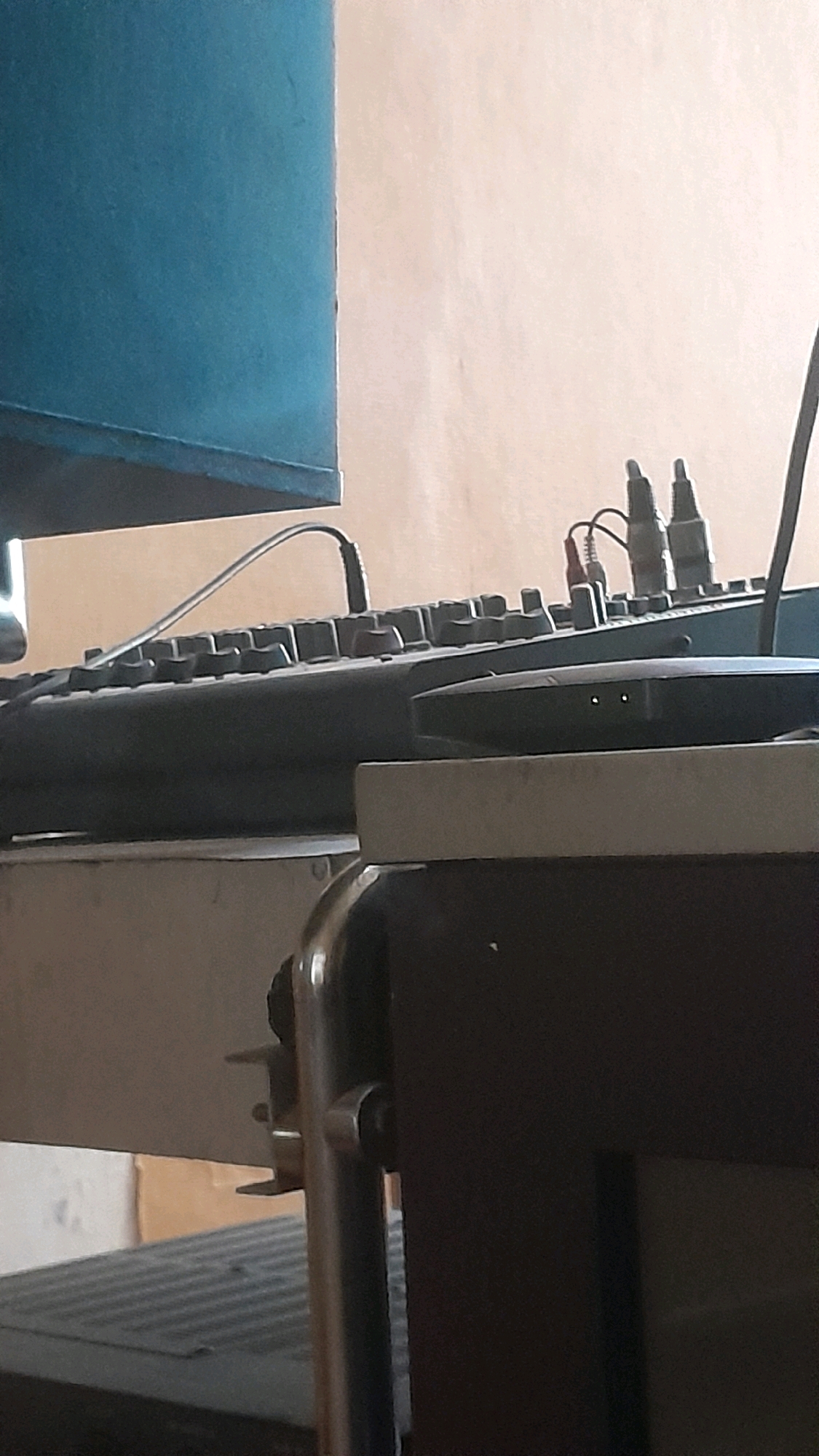 The image shows a close-up of a desk setup, likely in an office or home studio environment, with electronic equipment. In the foreground, a black electronic device with two small indicator lights is visible. Behind it, a grey keyboard with many keys is partially obscured. A thin grey cable arches over the keyboard. Further back, a dark teal-colored electronic device, possibly a mixing board or audio interface, has several knobs and connectors, including two prominent black connectors with red and white wires attached. The background is a neutral, light brown wall. The bottom part of the image shows a glimpse of another electronic device with a textured surface, and a metallic support structure. The scene is indoors, with artificial lighting. There are no people visible in the image. The location in Madiun, Indonesia, cannot be determined from the visual information provided.