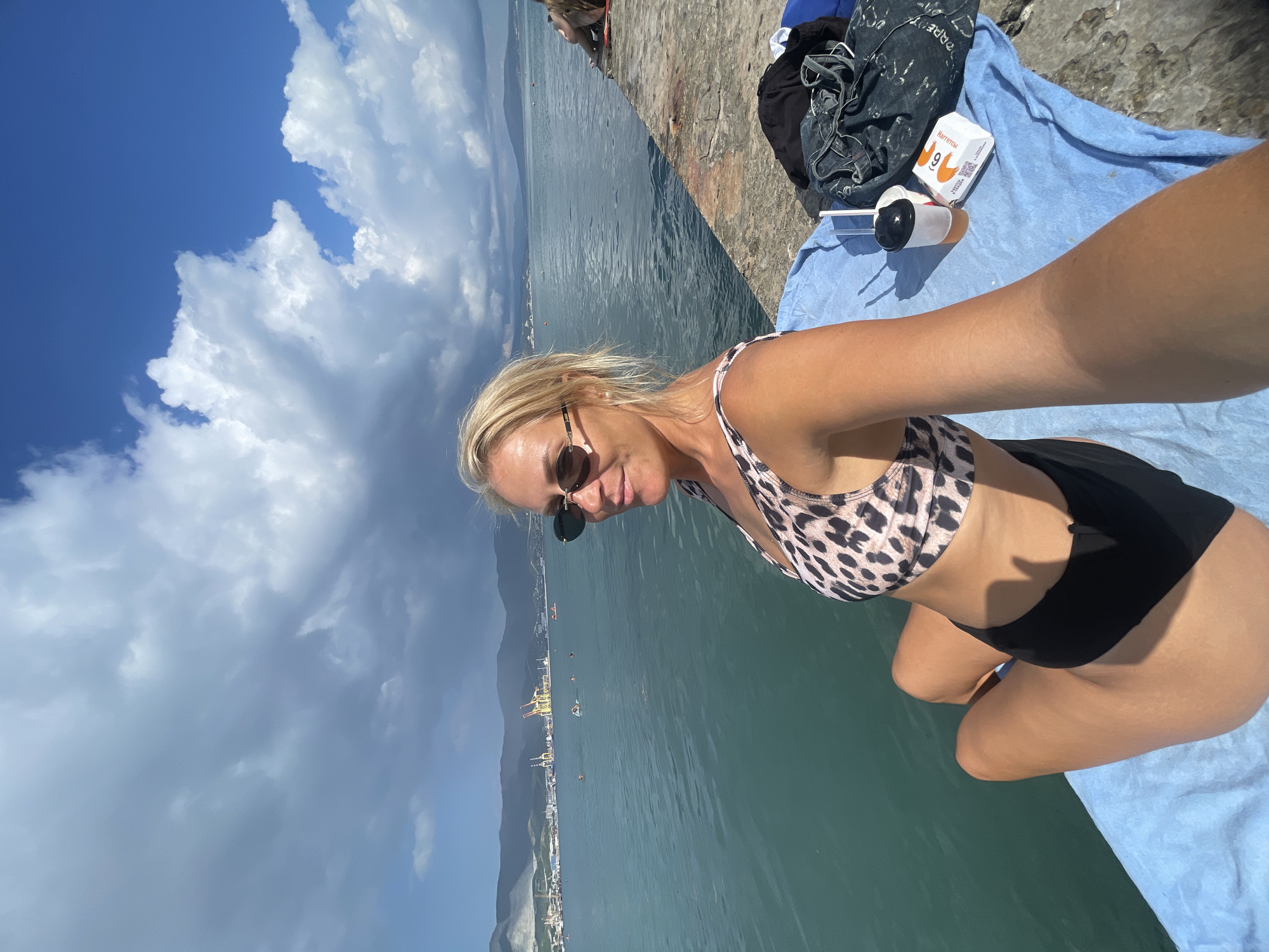 In Novorossiysk, Russia, a blonde woman in a leopard-print bikini top and black bottoms sits on a blue towel on a concrete pier. She is wearing sunglasses and has her arm extended, holding the camera for a selfie. To her right, on the towel, are a paper cup with a straw, a small white box with an orange logo, and a dark denim garment. The sea is calm and a light blue-green, with distant mountains and industrial cranes visible on the horizon under a partly cloudy sky. Several people are swimming in the water. The overall scene depicts a woman enjoying a day at the seaside.