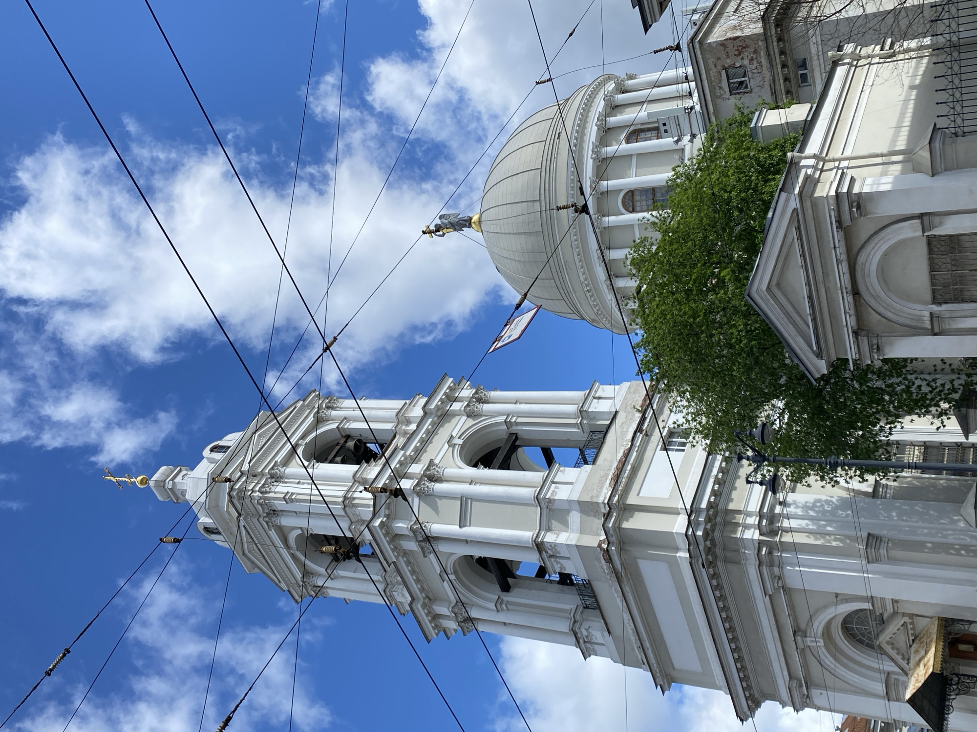 The image shows a low-angle view of architectural structures in Saint Petersburg, Russia, against a blue sky with scattered white clouds. A tall, multi-tiered, white bell tower with arched openings and a gold cross at its peak stands prominently. Adjacent to the bell tower is a large, domed building with columns and a statue on top. Power lines crisscross the sky in the foreground. Green foliage is visible near the domed building. The overall scene suggests a historic urban landscape in Saint Petersburg.