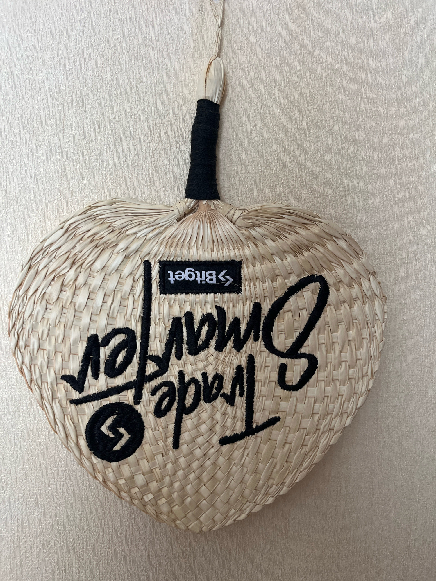 A woven straw fan hangs on a textured, light-colored wall. The fan has a heart-like shape with a black handle wrapped in thread. Black embroidered text on the fan reads "Trade" in large script at the bottom and "Bitget" in a smaller rectangular patch above it. A black circular logo with a stylized lightning bolt is embroidered below the word "Trade." The fan appears to be a decorative item. The location is Dzoraghbyur, Armenia.