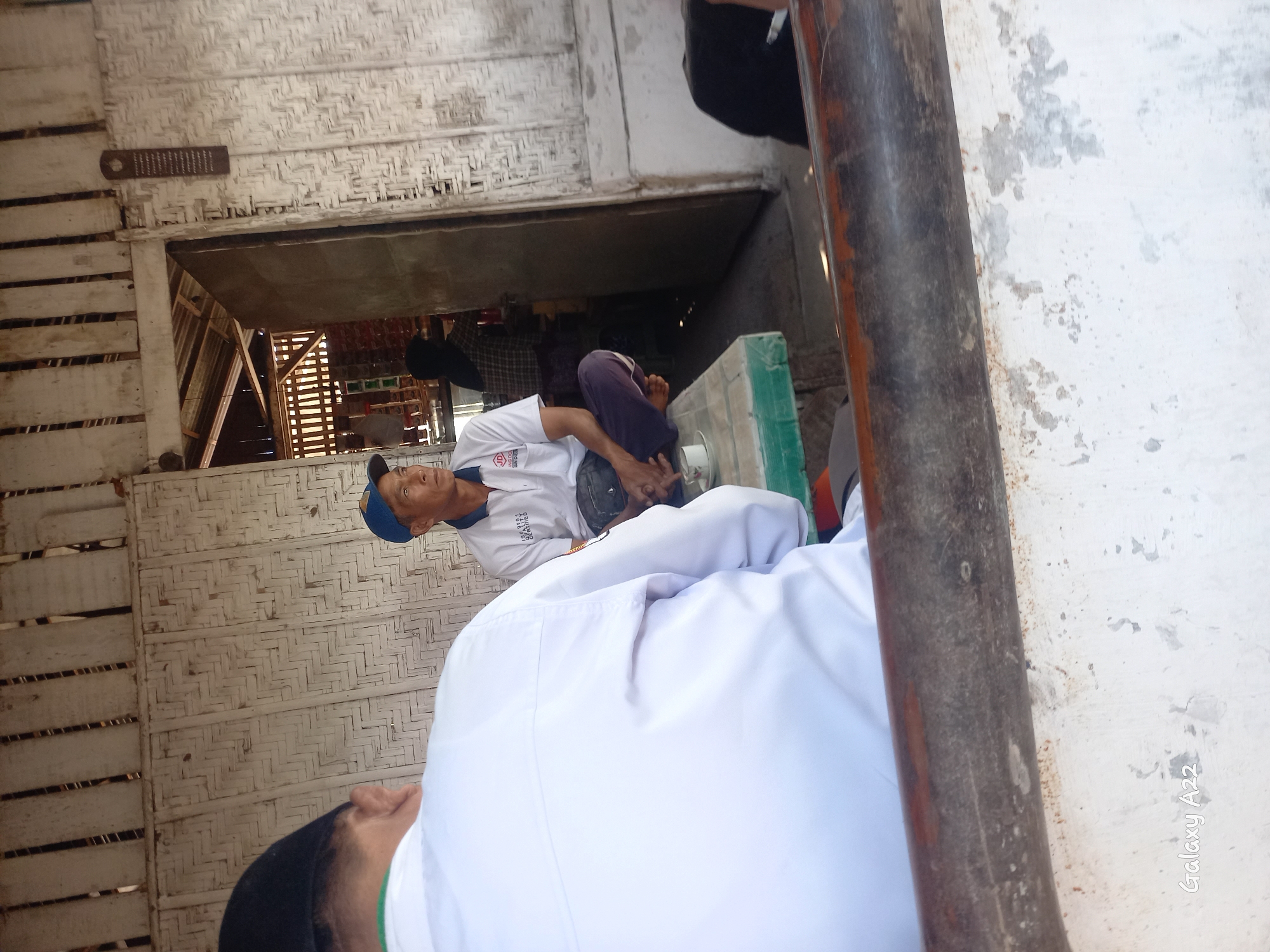 Two men are seated in what appears to be an open-air structure with woven bamboo walls and a wooden ceiling. The man on the left is partially visible from behind, wearing a white shirt and a black cap. He is facing away from the camera. The man on the right is seated on a green table, wearing a white polo shirt with a logo and a blue cap. He is looking to his right. A cup of liquid sits on the table in front of him. In the background, an open doorway reveals shelves stocked with goods, suggesting this might be a shop or stall. Another person, wearing a headscarf, is visible behind the counter. The overall environment appears rustic and informal. The image shows the "Galaxy A22" watermark in the bottom left corner. This scene is located in Panji Kidul, Indonesia.