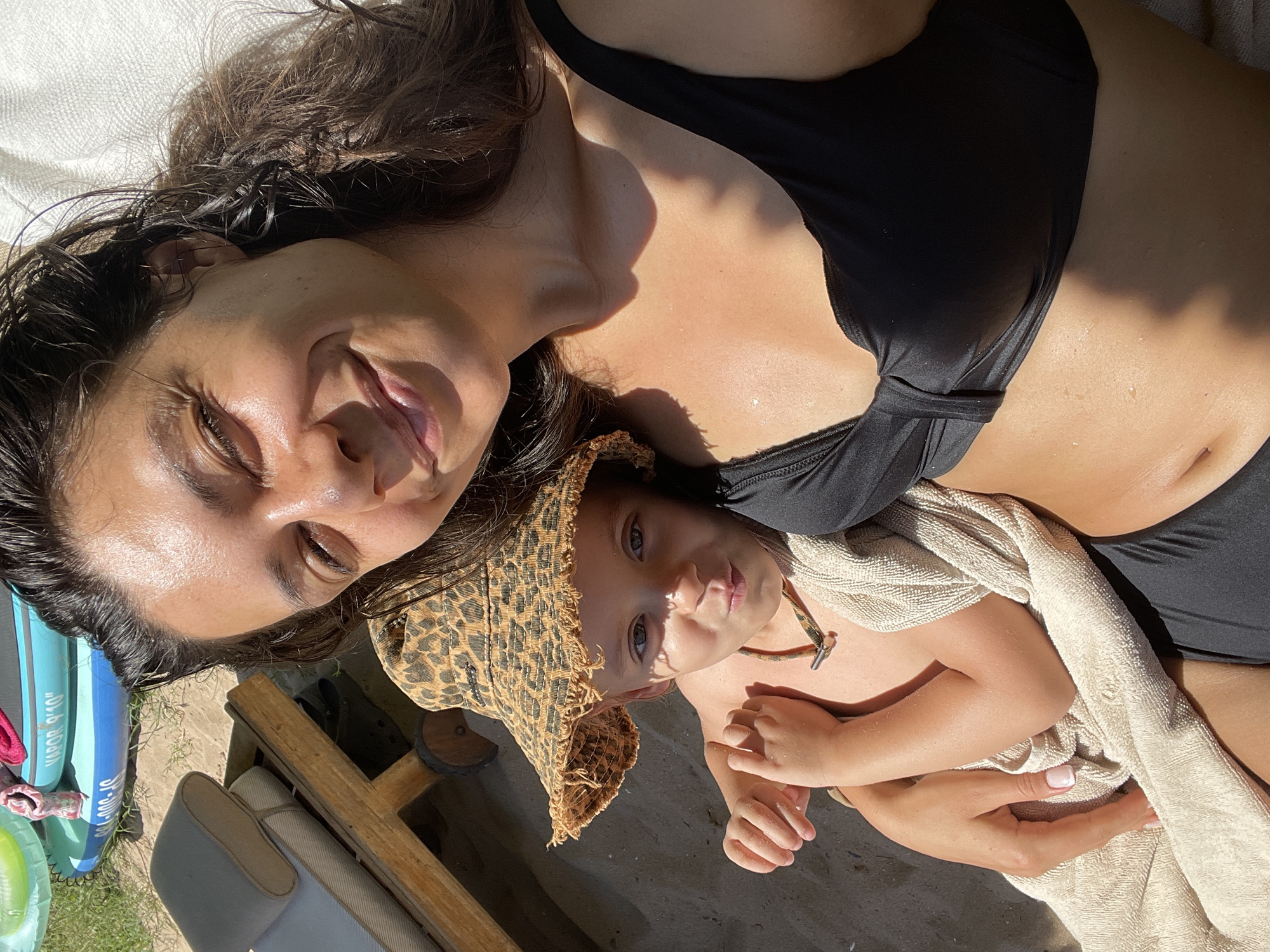 This media file depicts a close-up, overhead shot of two individuals, an adult and a child, lying down outdoors. The adult, with dark, wet hair and fair skin, is wearing a black bikini top. They appear to be smiling with their eyes closed or partially open. The child, wearing a leopard-print wide-brimmed hat and wrapped in a beige towel, is looking directly at the camera with a neutral expression. Their hands are visible, one resting on the adult's chest. The overall scene suggests a casual, relaxed moment, possibly at a beach or by a pool, given the presence of inflatable objects in the background. The lighting indicates it is daytime, with harsh sunlight casting shadows. The presence of sand and grass in the background provides further context for an outdoor setting. The visible text on the inflatable objects is partially obscured but includes "VAPOR 9'10"". The image conveys a sense of warmth and connection between the two subjects. The location context is Chernomorets, Bulgaria.