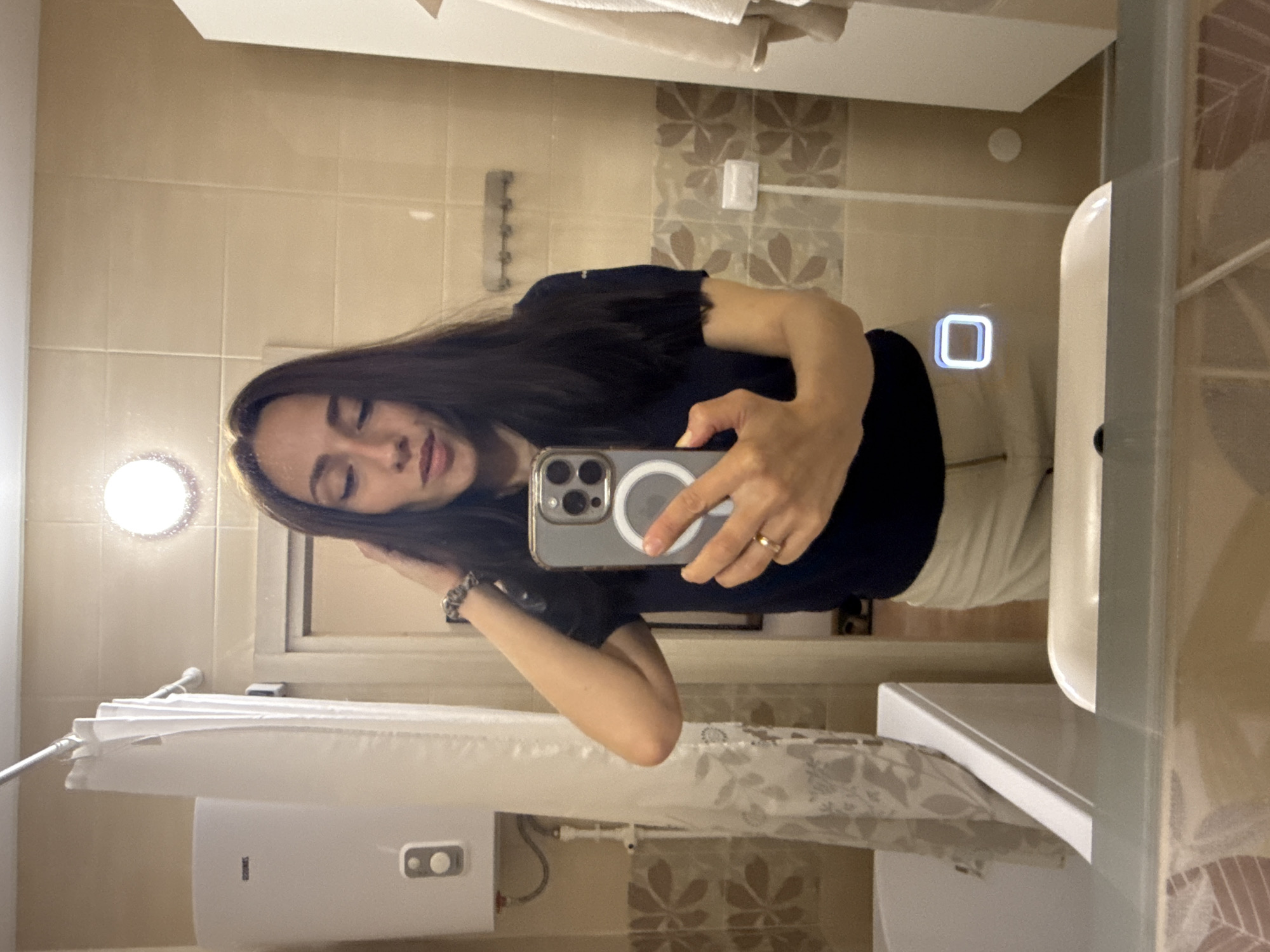 A dark-haired woman takes a selfie in a bathroom with cream-colored tile walls. Her phone is an iPhone with a silver case and a magnetic charging ring. She wears a black shirt and white pants. A light is visible in the upper left corner. A towel rack is on the wall above the sink. A water heater is visible to the left of the image. The location cannot be determined from the image, nor can it be reliably connected to Saint Petersburg, Russia.
