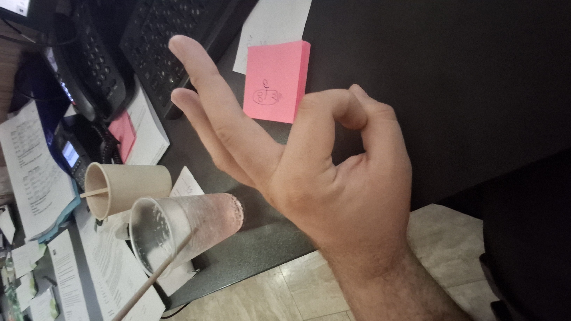 The image shows a close-up of a person's hand making an "OK" sign over a stack of pink sticky notes. On the sticky notes, there is a crude drawing of a ghost. The scene appears to be an office or desk area, with papers, a telephone, and a keyboard visible in the background. The floor tiling suggests it could be a commercial or office space. The image does not provide any specific information about the city of Nesebar, Bulgaria.