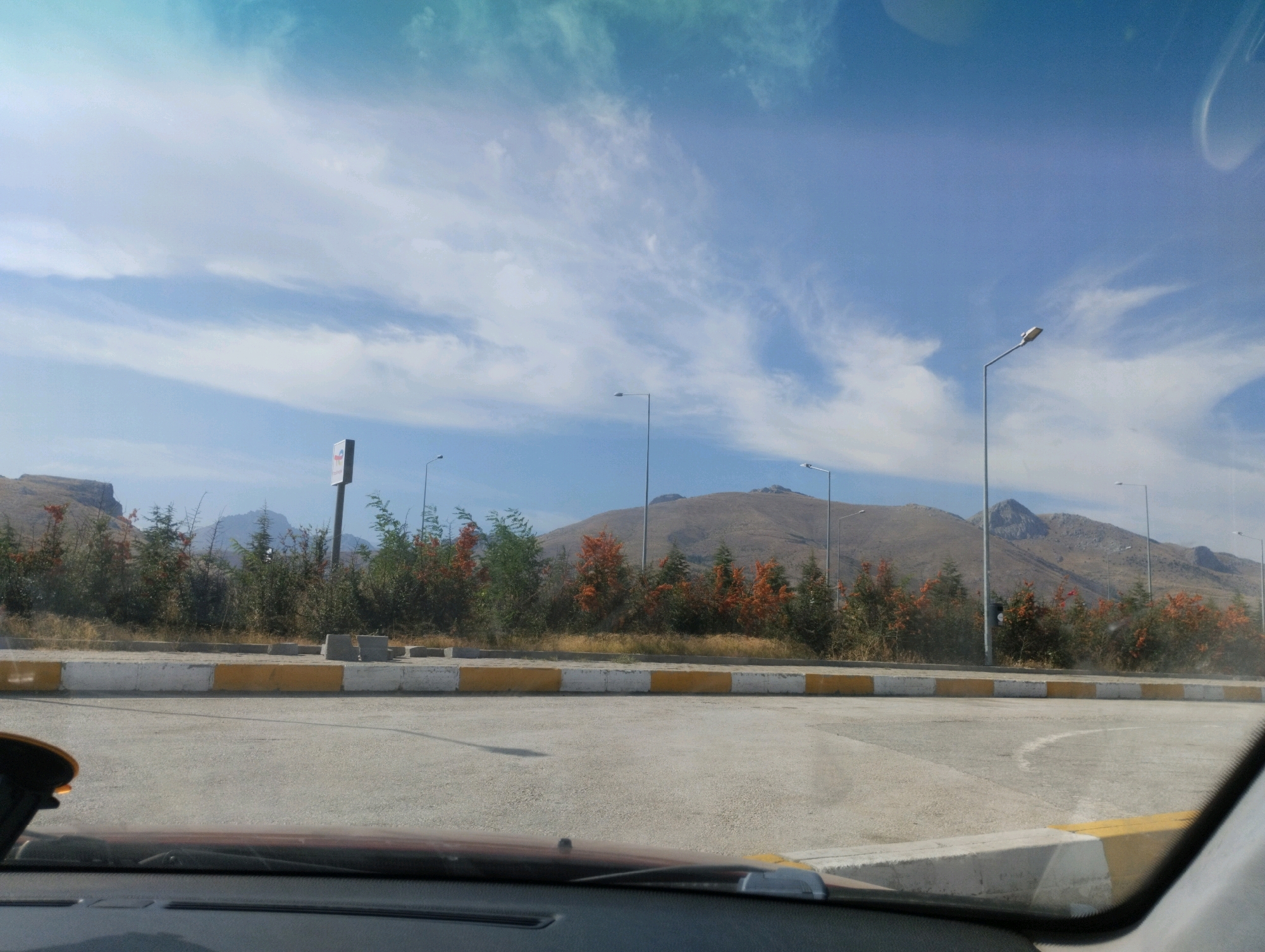 A daytime, outdoor, eye-level, medium shot captures a road with a view of a natural landscape. The foreground shows the interior of a vehicle, including the dashboard and part of the windshield. The road itself is paved with asphalt and marked with white and yellow lines on the curb. Behind the road, a row of trees with green and orange foliage runs horizontally, with some dry grass and sparse vegetation. In the background, a large, barren mountain range under a clear blue sky with scattered white clouds dominates the scene. Several modern streetlights are visible along the roadside, extending into the distance. A directional sign is positioned on the left side of the road. The scene appears to be a roadside area in Ulukışla, Turkey, likely an exit or entrance to a highway or a scenic viewpoint.
