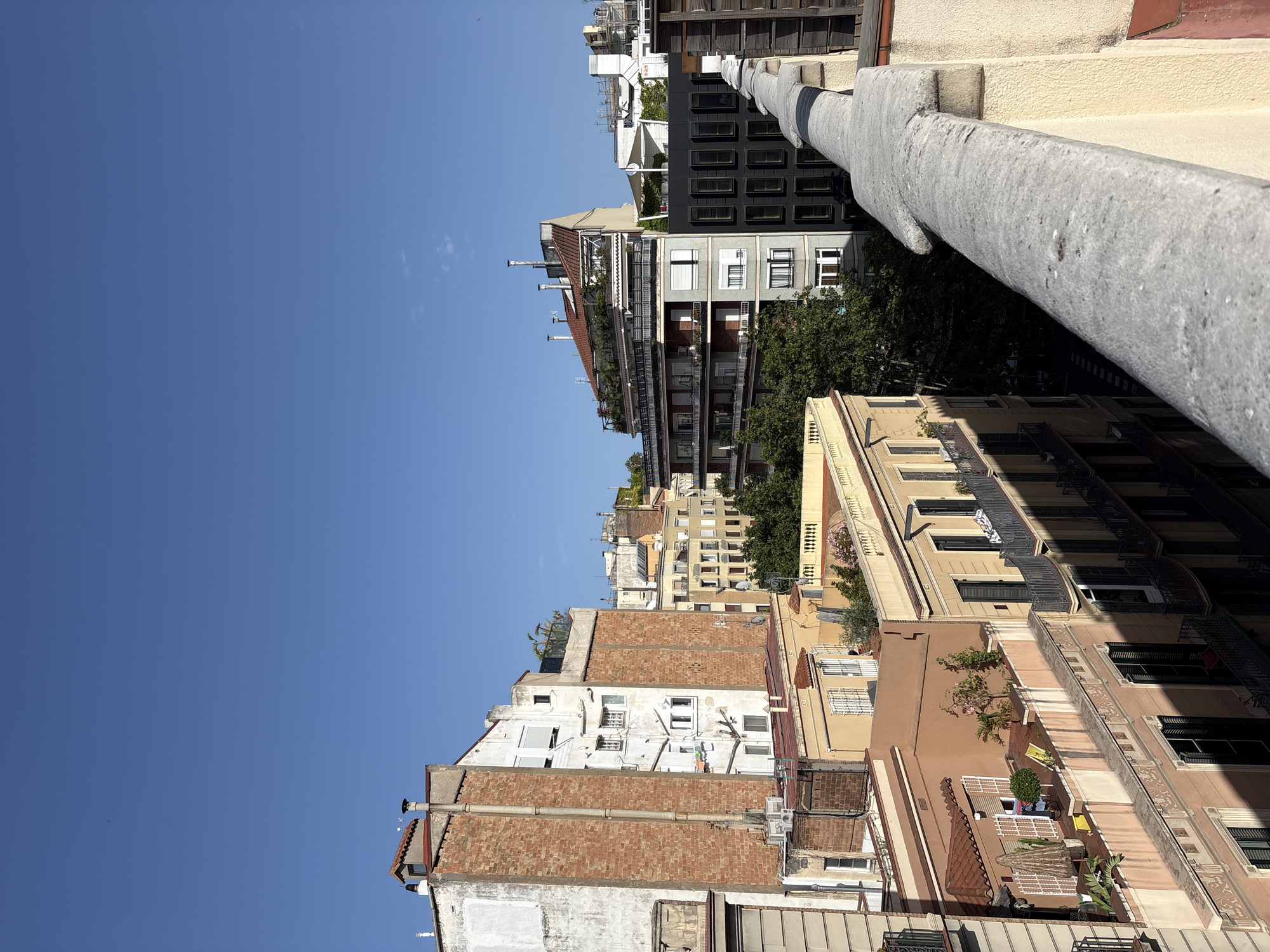 This is a high-angle photograph of a city skyline, likely Barcelona, Spain, under a clear blue sky. The scene is dominated by various multi-story buildings with distinct architectural styles. In the foreground, a textured, concrete railing runs diagonally across the lower right portion of the frame, suggesting the viewpoint is from a rooftop or balcony.
To the left, the side of a building with a red brick facade and white plaster accents is visible. It has several windows and a tall, gray vertical pipe running along its exterior. Adjacent to this is a cream-colored building with multiple floors and white-shuttered windows.
Further into the middle ground and to the right, a collection of buildings with different colors and designs are arranged. One building has a distinctive terracotta-tiled roof with a rooftop garden featuring lush greenery and potted plants. Another prominent structure in the mid-right is a dark gray building with a grid-like pattern of rectangular windows. Between these buildings, dense green trees are visible, indicating the presence of courtyards or larger green spaces within the urban environment.
The buildings exhibit details such as balconies with railings, air conditioning units, chimneys, and decorative architectural elements. The lighting suggests it is daytime, possibly late morning or early afternoon, with strong sunlight casting shadows. There are no people or animals clearly visible in the image. The overall impression is one of a dense urban landscape with a mix of older and newer architectural styles.