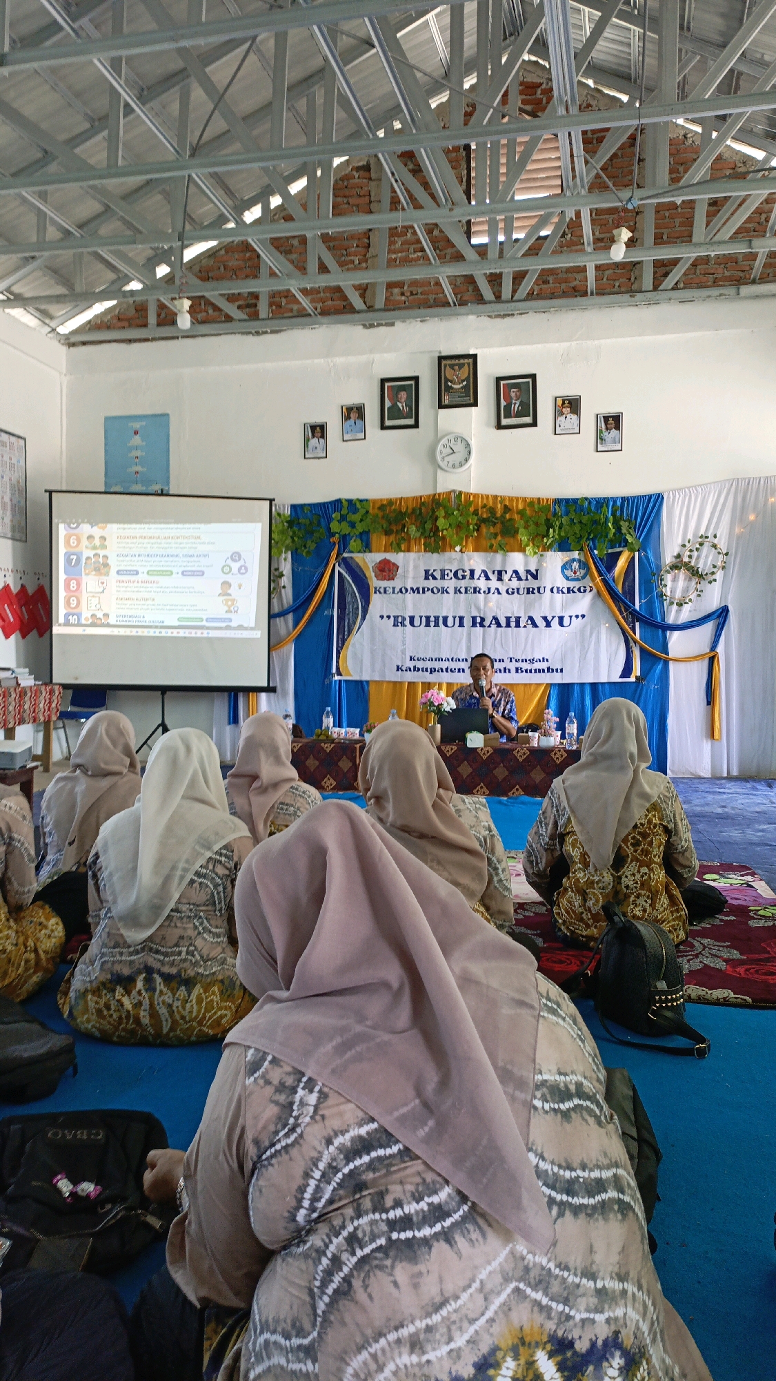 The image depicts an indoor gathering in Salimuran, Indonesia, likely a meeting or workshop for teachers. In the foreground, several individuals wearing headscarves and patterned clothing are seated on the floor, facing a projection screen and a stage area. The projection screen displays information in Indonesian, possibly related to an educational topic. On the stage, a man in a patterned shirt is speaking, appearing to be a presenter or facilitator. Behind him, a banner with Indonesian text announces "KEGIATAN KELOMPOK KERJA GURU (KKG)" which translates to "Teacher Working Group Activity" and the subtitle ""RUHUI RAHAYU"". The room has exposed brick walls and a metal ceiling structure. Pictures of notable figures and a clock are visible on the wall above the stage. The overall atmosphere suggests a formal yet communal educational event.