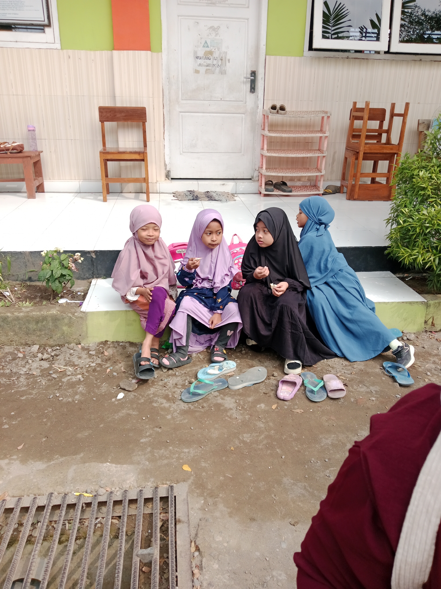 Four young girls wearing hijabs are sitting on a raised concrete ledge outdoors. The girl on the far left wears a light purple hijab and a purple and white patterned outfit. Next to her, a girl in a darker purple hijab and a dark blue and patterned dress holds a small white object. The third girl from the left is dressed in black, with a black hijab, and is also holding a small white object. The girl on the far right wears a light blue hijab and a matching blue dress. Scattered around them on the ground are various pairs of sandals and flip-flops in different colors. The background features a building with a white door, a wooden chair, a small pink shelf unit, and some greenery. The ground is a mix of dirt and gravel. The image appears to be taken during the daytime. This scene is consistent with an outdoor area in Kediri City, Indonesia, potentially near a school or community building.
