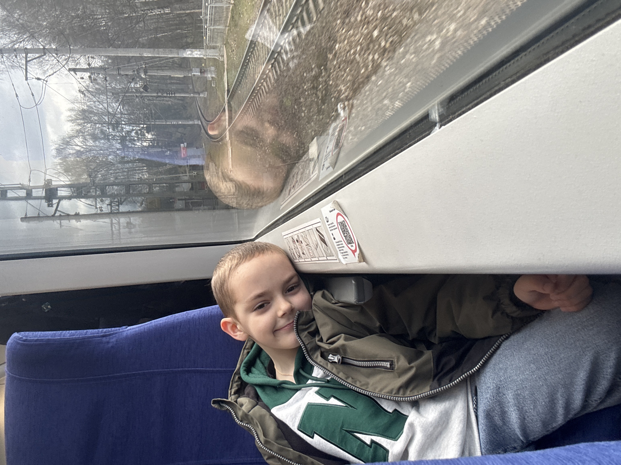 A boy is sitting inside a train car. He is wearing a green and white sweatshirt with a large "N" on it, a brown jacket, and jeans. He is smiling. A blue seat is visible. Outside the train window, a railway track, trees, and overhead power lines are visible. The exact location cannot be determined from the image alone.