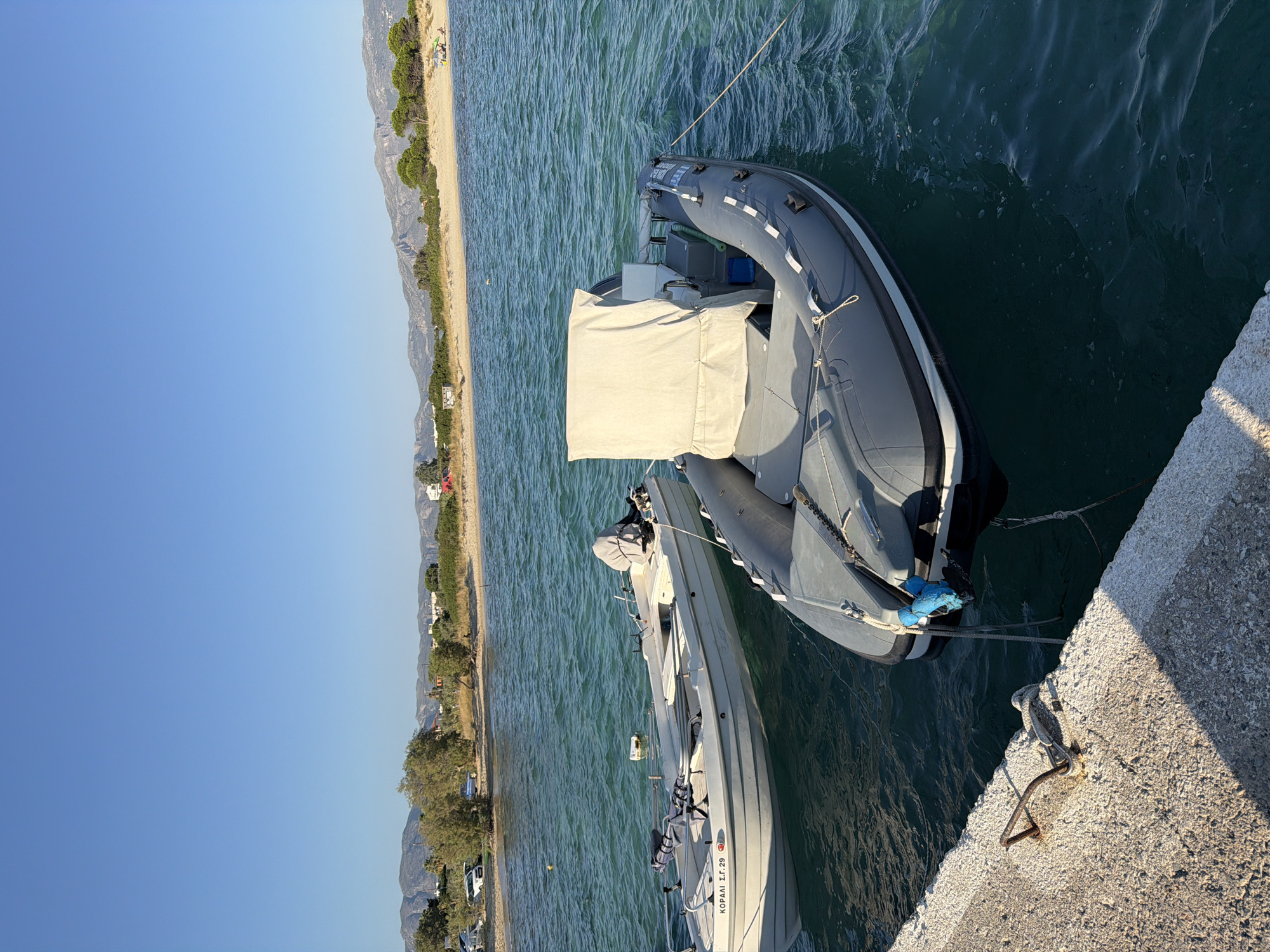 Two inflatable boats are docked at a concrete pier on a sunny day. The boat in the foreground is gray and has a beige cover over part of its interior. The boat in the background is white with some equipment visible, including seats and a trolling motor. The water is a clear blue-green and appears calm. In the distance, a sandy beach is visible with trees and hills beyond. The sky is clear and blue. There are no people visible in the image. The location is the seaside of Viglafia, Greece.