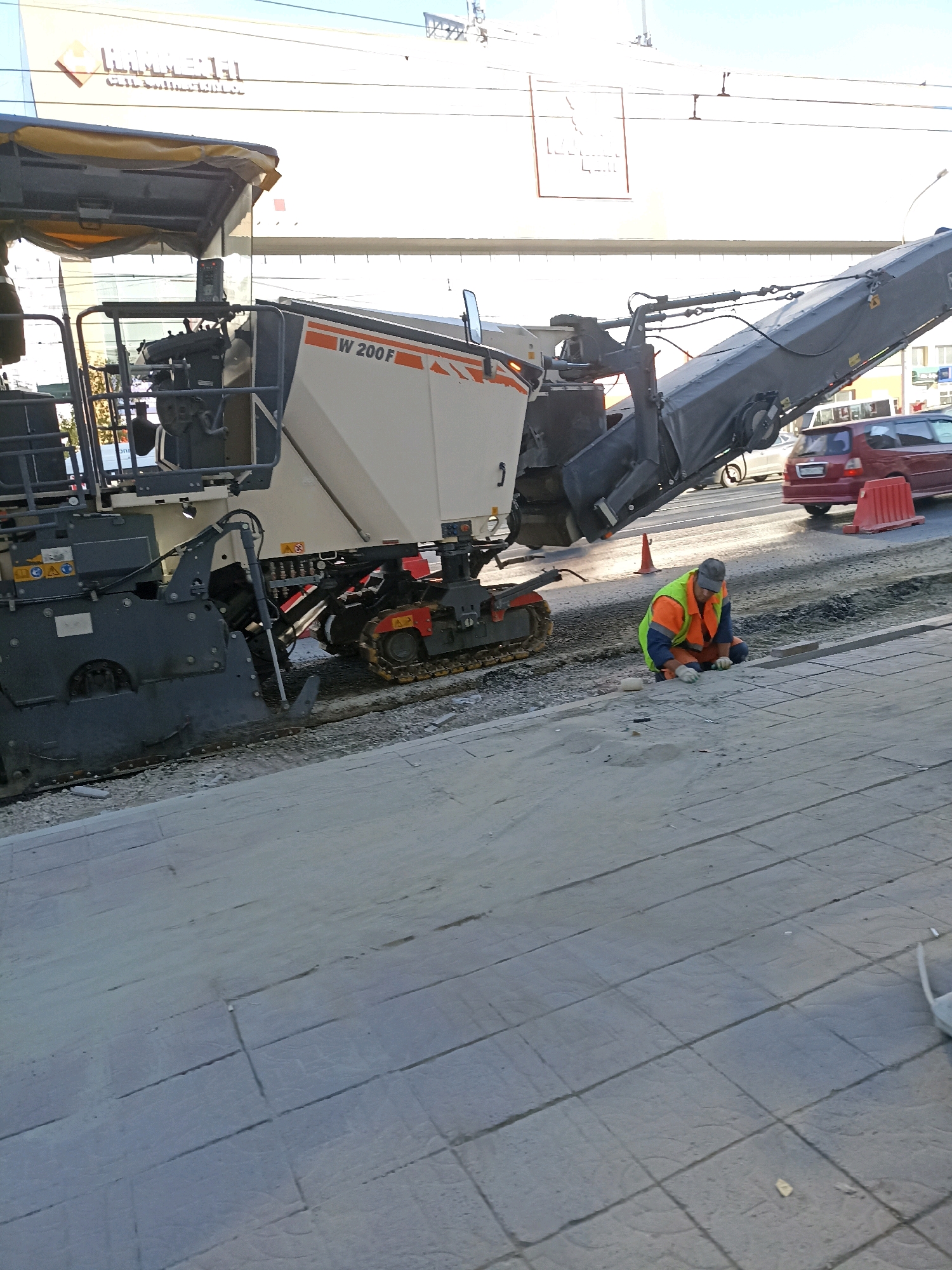 In an outdoor urban setting, a road milling machine, labeled "W 200F," is positioned on a construction site. A worker wearing a high-visibility orange vest over a green shirt and blue pants, along with white gloves and a gray cap, is kneeling on the ground near the milling machine. He appears to be working with some materials on the paved surface. In the background, a red car is visible on a road, and construction elements like barriers and traffic cones are present. Overhead, power lines and the facade of a building with signage that appears to read "LINOMEL" are present. The ground is paved with light-colored rectangular tiles.
