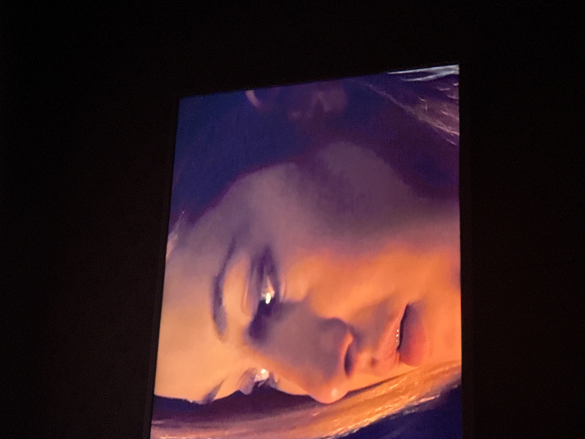 The image shows a close-up of a woman's face displayed on a screen, likely a TV or monitor. The face is angled, with a focus on the eye, nose, and lips. The lighting is warm, giving the skin an orange hue. The background surrounding the screen is black. There is no discernible information to place the image within Mytishchi, Russia, or any other specific location.
