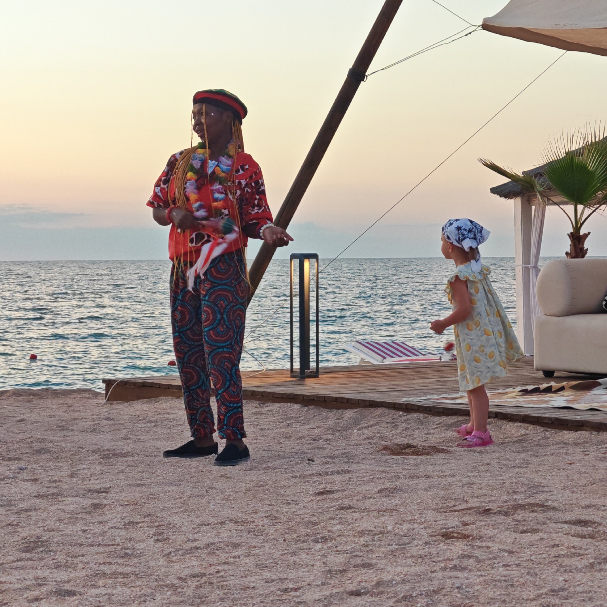 This image depicts an outdoor scene at the beachfront of Мысовское сельское поселение, Ukraine, during what appears to be sunset or dawn. The sky is a pale orange and yellow, with some muted blue clouds near the horizon. The sea is calm with gentle waves.
In the foreground, on a sandy and shell-covered beach, stands an adult dressed in a vibrantly patterned red shirt and matching pants. The shirt has a colorful print with floral and abstract designs. They are wearing a black beret with red and green accents and have blonde braids hanging down. The adult is holding what looks like a fan or a collection of feathers in their right hand and is looking to the side.
To the right of the adult, a young child is standing on a wooden deck. The child is wearing a light-colored dress adorned with yellow lemon prints and a patterned headscarf. They are facing away from the viewer, looking out towards the sea.
Behind the figures, a wooden deck extends towards the sea. On the deck, there is a modern black outdoor lamp with a warm light. Further back, a white tent-like structure with a thatched roof is visible, along with a plush white sofa and a palm tree. A beach chair with red and white stripes is also present on the deck. The overall atmosphere suggests a relaxed, possibly resort-like, setting.