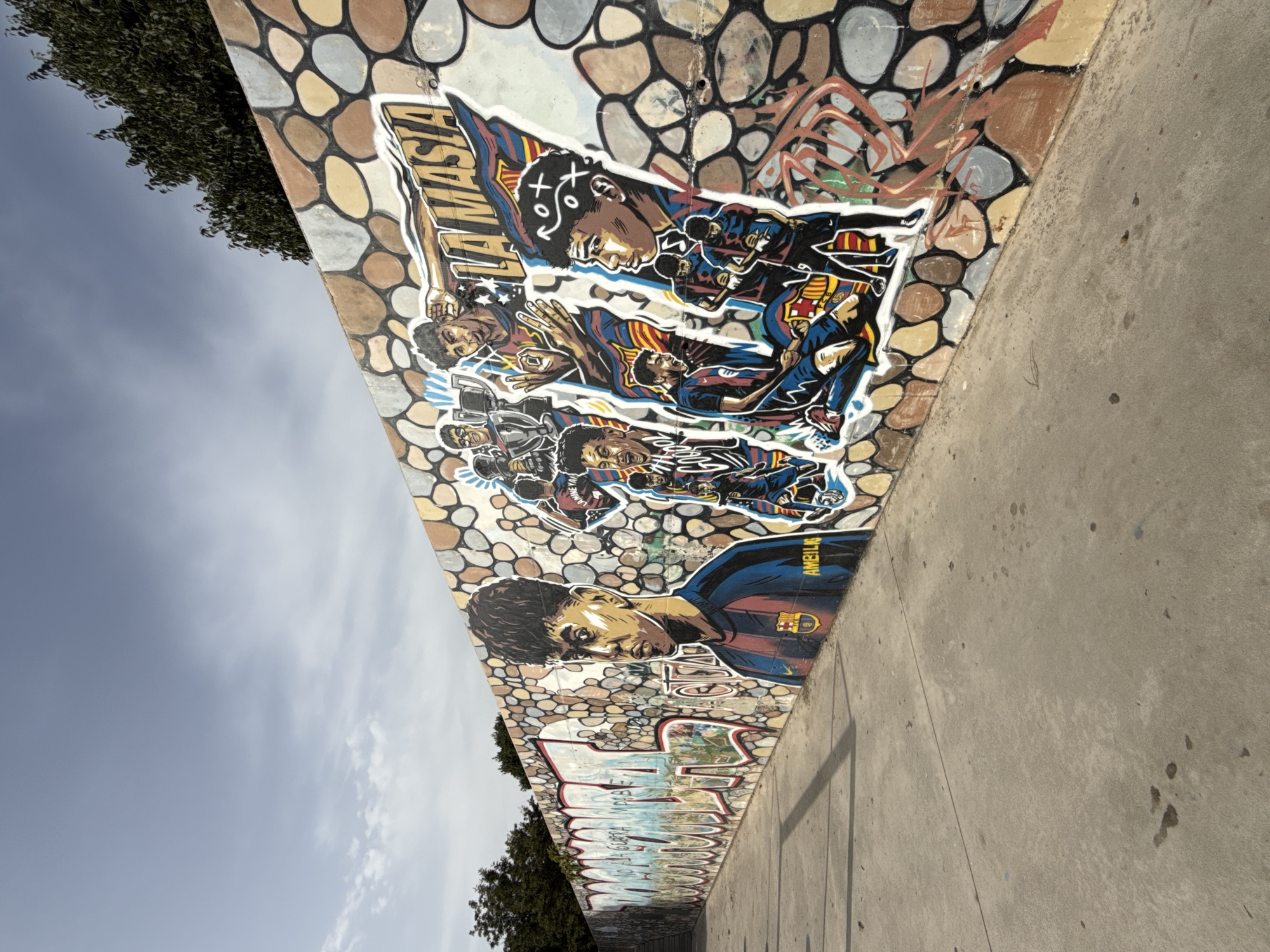 In Mataró, Spain, on a wall adorned with a mosaic of colorful stones, a vibrant graffiti mural depicts several young men in soccer uniforms. The central figure, a young man with a serious expression, wears a blue and red striped jersey with a FC Barcelona crest. To his right, a dynamic composition features other young men celebrating, one holding a trophy and another making a gesture with his hand. Text in Spanish, including "LA MASIA," the name of FC Barcelona's youth academy, is integrated into the artwork. The mural stretches along a concrete walkway, with trees visible in the background against a cloudy sky. The overall scene captures a spirited and artistic representation of soccer culture.