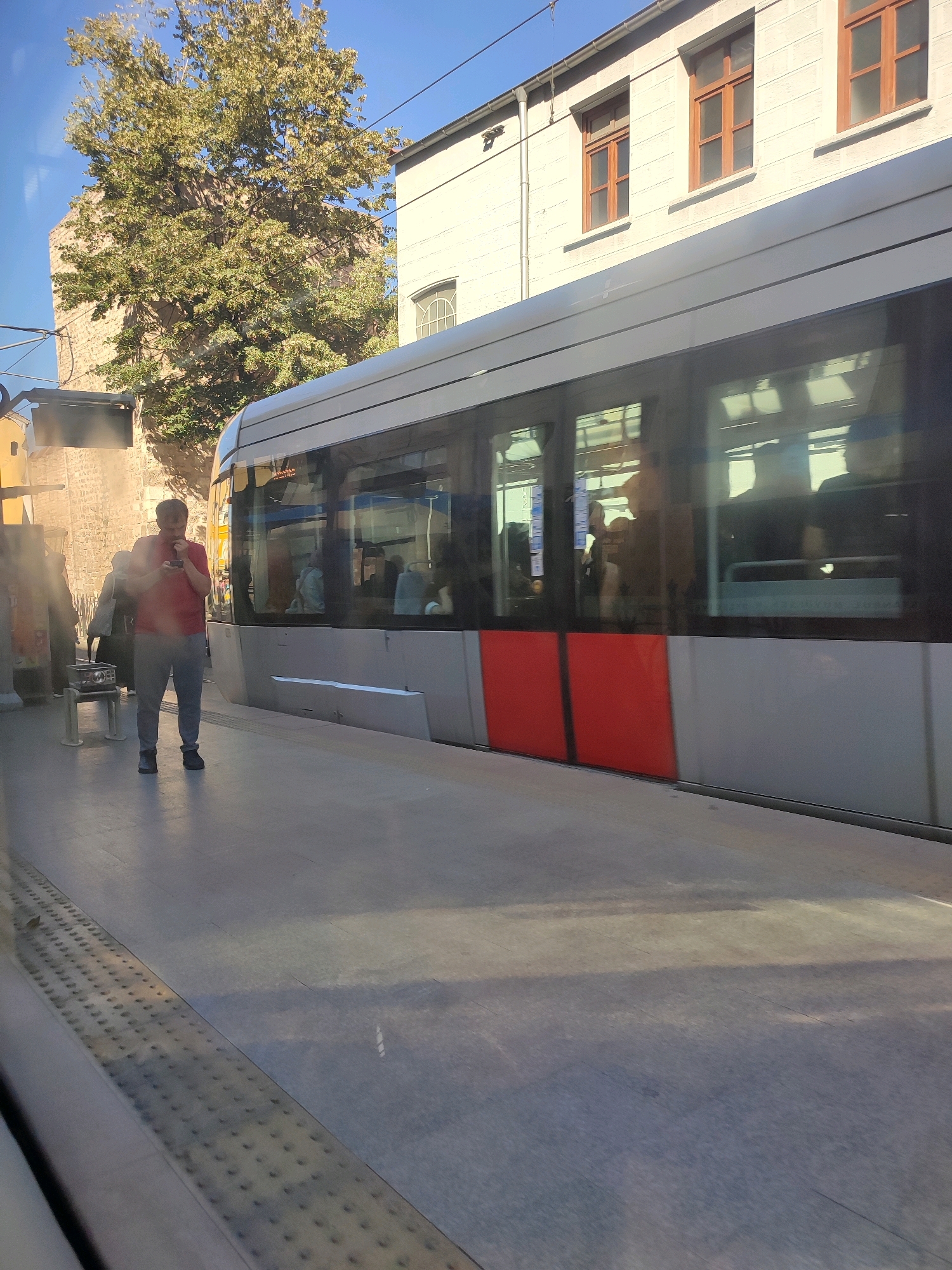 A man wearing a red shirt and grey pants is standing on a train platform in Istanbul, Turkey, looking at his phone. He is wearing dark shoes and carrying a dark bag over his shoulder. Next to him is a white bench with a metal frame. Behind him, a modern tram with grey and red panels is stopped at the platform. The tram has large windows that reflect the surroundings, showing blurred images of people inside. To the left of the man, there is a low wall with some graffiti and a building with a stone facade. In the background, a tall, leafy tree stands against a clear blue sky, and beyond that, a building with a white facade and brown-framed windows. The platform is paved with grey concrete tiles, and there are tactile paving strips along the edge. The scene is illuminated by bright sunlight, casting shadows on the platform.