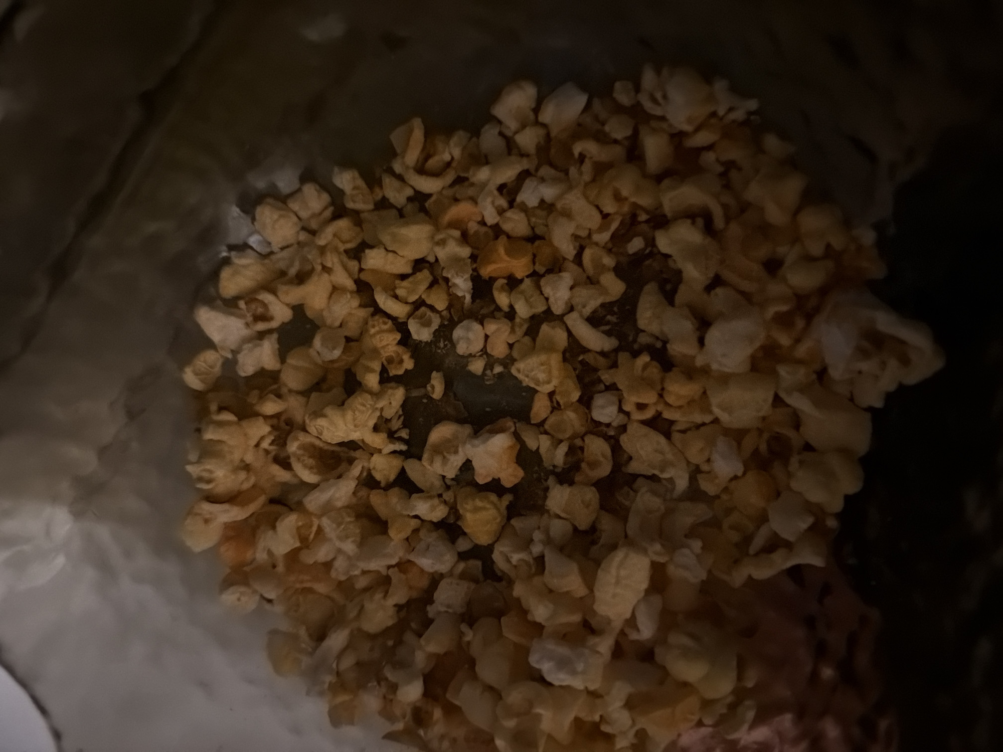 The image shows a close-up, overhead view of a pile of popcorn. The popcorn is mostly light yellow and white, with some pieces appearing slightly orange. The popcorn is irregular in shape and size, with some pieces appearing puffed and others more flattened. The popcorn is contained within a dark, possibly paper, bag or container. The lighting is dim, casting shadows and highlighting the texture of the popcorn. There are no people or other objects visible in the image. The image does not contain any information that would allow for the identification of a specific location in Kyiv, Ukraine, or any other city.