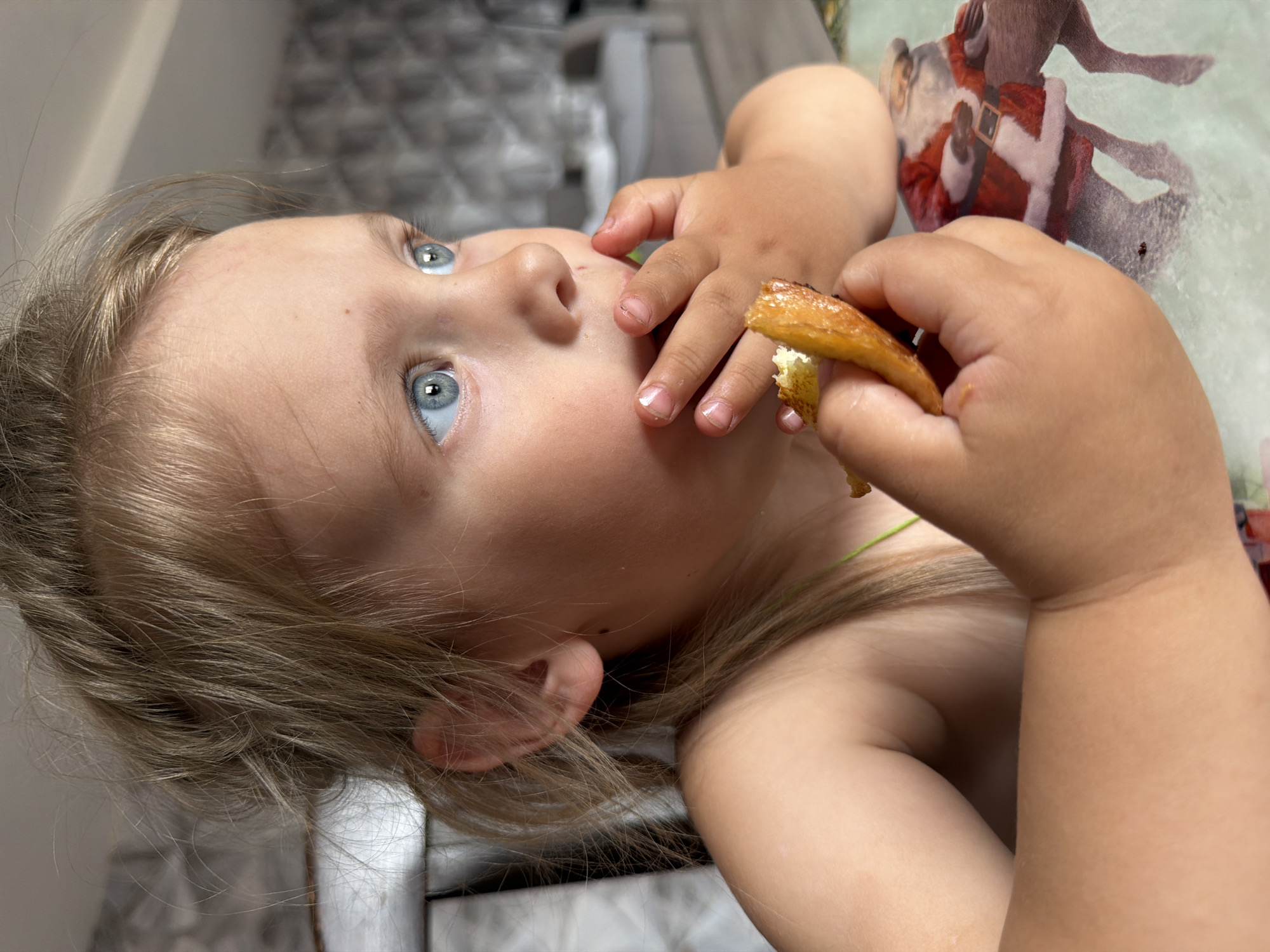 This close-up image, set in Chaikovsky, Russia, features a young child, likely a toddler, with light blonde hair and striking blue eyes. The child's head is tilted back, looking upwards and to the left with an expression of focused curiosity.
The main activity depicted is the child eating. Both small hands grasp a piece of golden-brown food, possibly a fried pastry, bread, or pancake, from which a bite has been taken, revealing a lighter interior. The child's fingers, with neatly trimmed nails, are visible around the food.
The setting appears to be indoors, likely a home, with a blurred background showing indistinct elements of a room, possibly including a light-colored tiled surface. A notable detail in the background is a visible illustration of Santa Claus on what appears to be a fabric surface, such as a blanket or tablecloth. A thin, light green string or strap is visible near the child's shoulder. The image is oriented sideways, suggesting the photo was taken from a low angle or that the child was lying down or held in a unique position. No text is visible in the image.