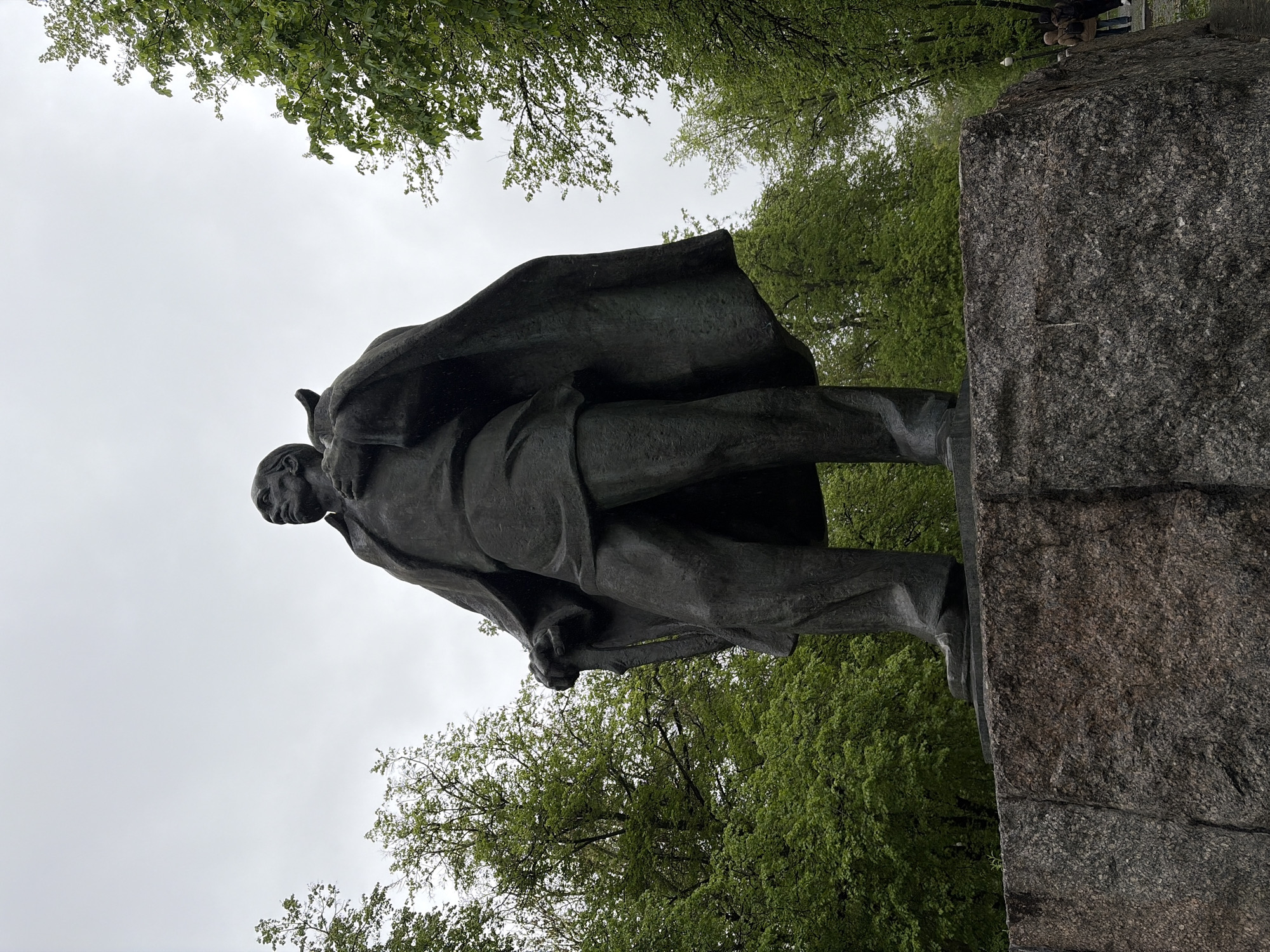 A low-angle shot shows a bronze statue of a man standing on a large, textured granite plinth. The man is dressed in a long coat or cape, and his head is turned slightly to the left. The surrounding environment includes trees with green foliage, seen from a low perspective, and a grey sky. The location appears to be a park or similar outdoor area, city of Unknown, Unknown.