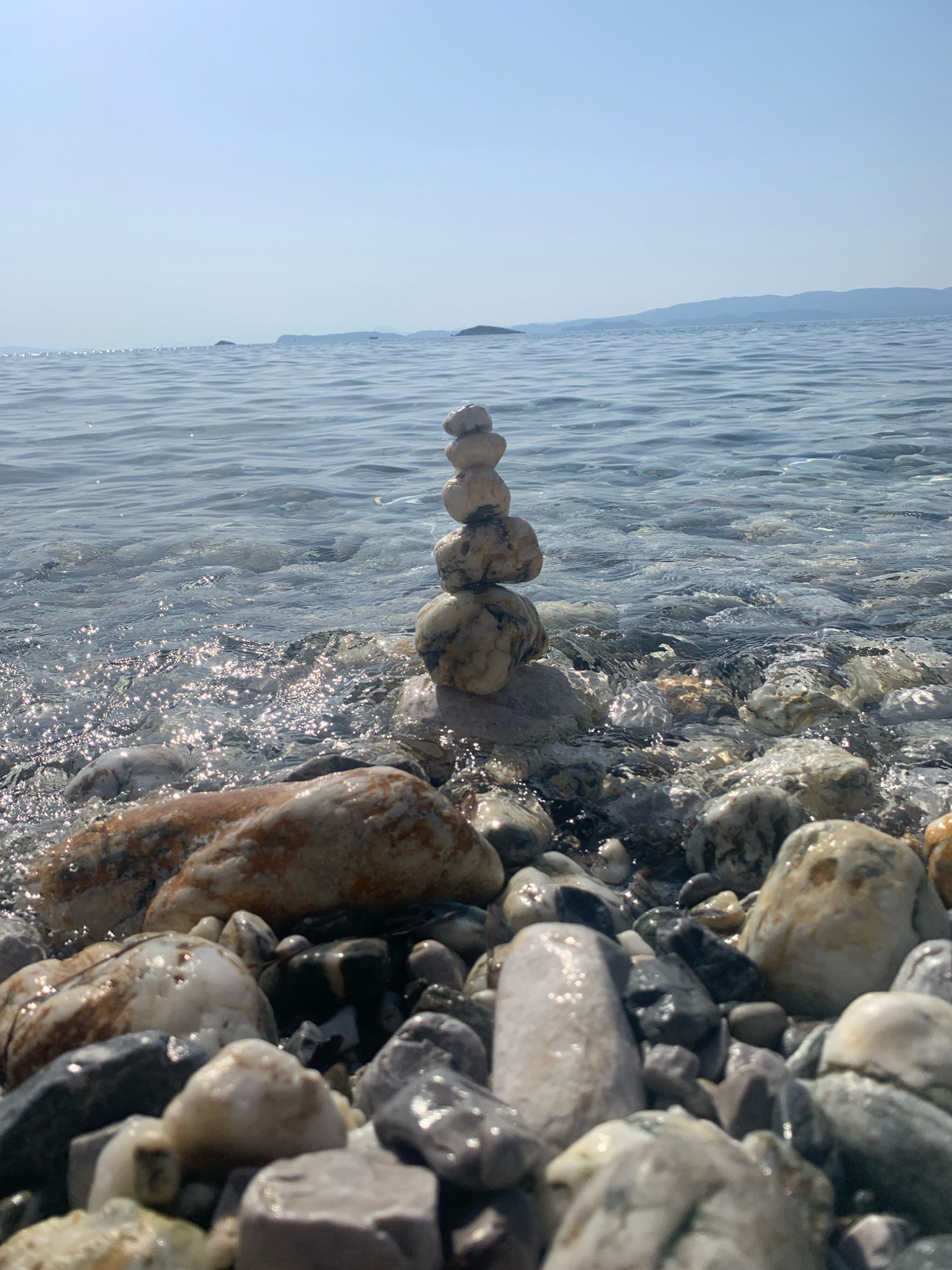 In Panormos, Greece, a stack of five smooth, weathered stones is precariously balanced on a pebble beach, with the clear, shallow water of the Aegean Sea gently lapping around its base. The sun reflects brightly off the water's surface, creating a shimmering effect. The beach itself is covered in a variety of smooth, rounded pebbles in shades of grey, white, and brown, some wet and dark, others dry and lighter. In the background, the calm, blue water stretches out to the horizon, meeting a hazy, light blue sky. Distant, low-lying hills or islands are visible on the horizon, their forms softened by the atmospheric perspective. There are no people or animals present in the scene.