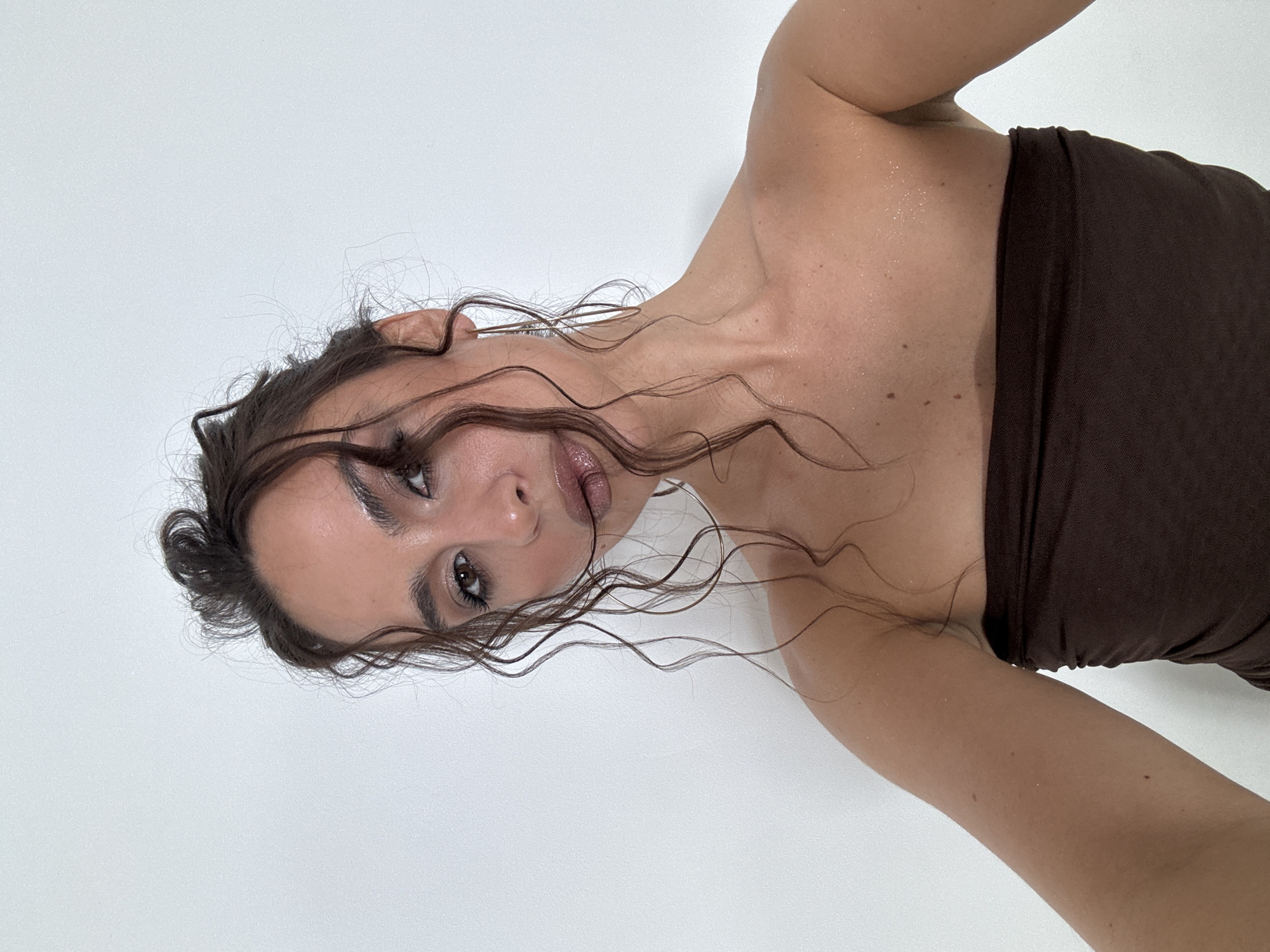 A light-skinned woman with brown hair pulled back in a messy bun, with strands framing her face, is oriented upside down against a white background. She is wearing a dark brown strapless top. Her skin appears to have a slightly glistening sheen, possibly due to sweat or moisturizer. Freckles are visible on her upper chest and shoulders. There are no indications of the location being Krasnoyarsk, Russia. The environment is a plain white backdrop.