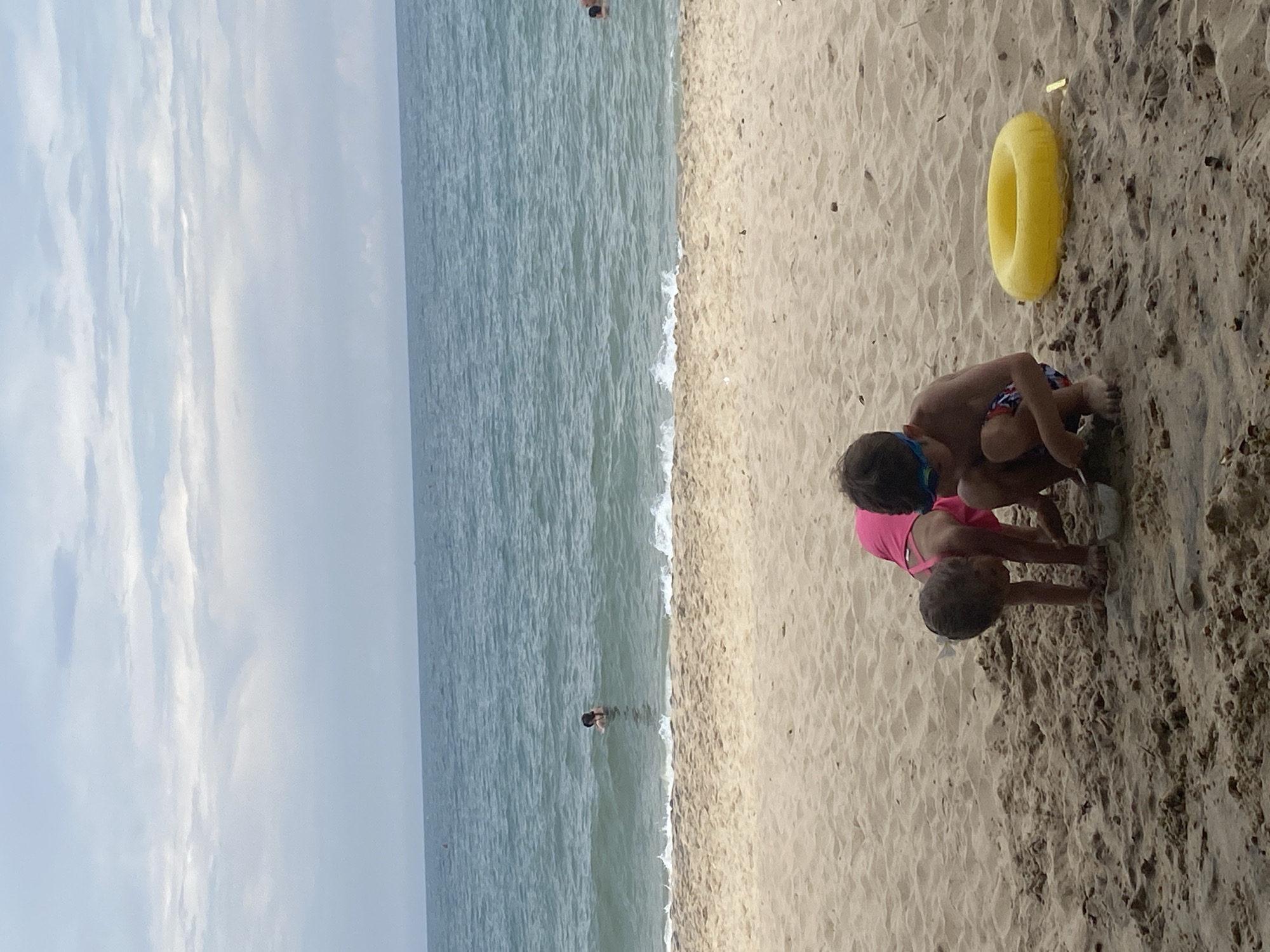 The image depicts a sandy beach with the ocean and sky in the background. Two children are playing in the sand in the foreground. A young boy wearing goggles and patterned swim trunks is crouched down. Next to him is a yellow inflatable ring. A younger child, wearing a pink swimsuit, is also crouched down, seemingly engaged in playing with the sand. In the ocean, several people are visible swimming. The sky is overcast with clouds. This scene is located on the beach in Phu Thuy Ward, Vietnam.