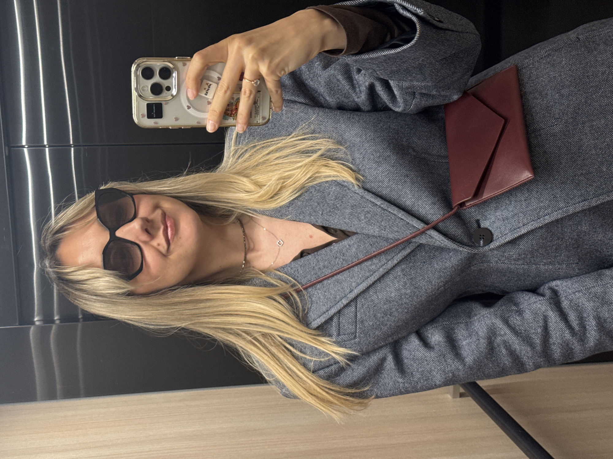 A woman is taking a selfie in what appears to be an elevator. She is wearing sunglasses, a gray blazer with a red purse slung across it, and a necklace. Her hair is blonde. She is holding a cell phone in her right hand, using it to take the picture. The background shows the interior of an elevator with metallic and wood paneling. It's impossible to confirm the location is Riga, Latvia, without additional information.