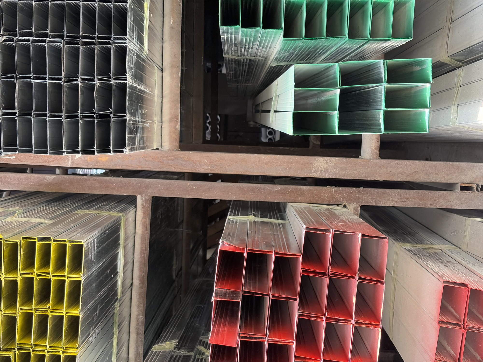 This image shows a storage rack filled with stacks of C-shaped metal framing pieces, commonly used in construction. The metal pieces are organized by color: yellow on the top left, gray on the top right, red in the middle left, and green on the bottom right. The racks themselves appear to be made of rusty metal, suggesting an industrial or warehouse setting. The lighting is dim, and the overall impression is one of a well-stocked but perhaps aging storage facility. The image is set in Novopokrovka, Kyrgyzstan.