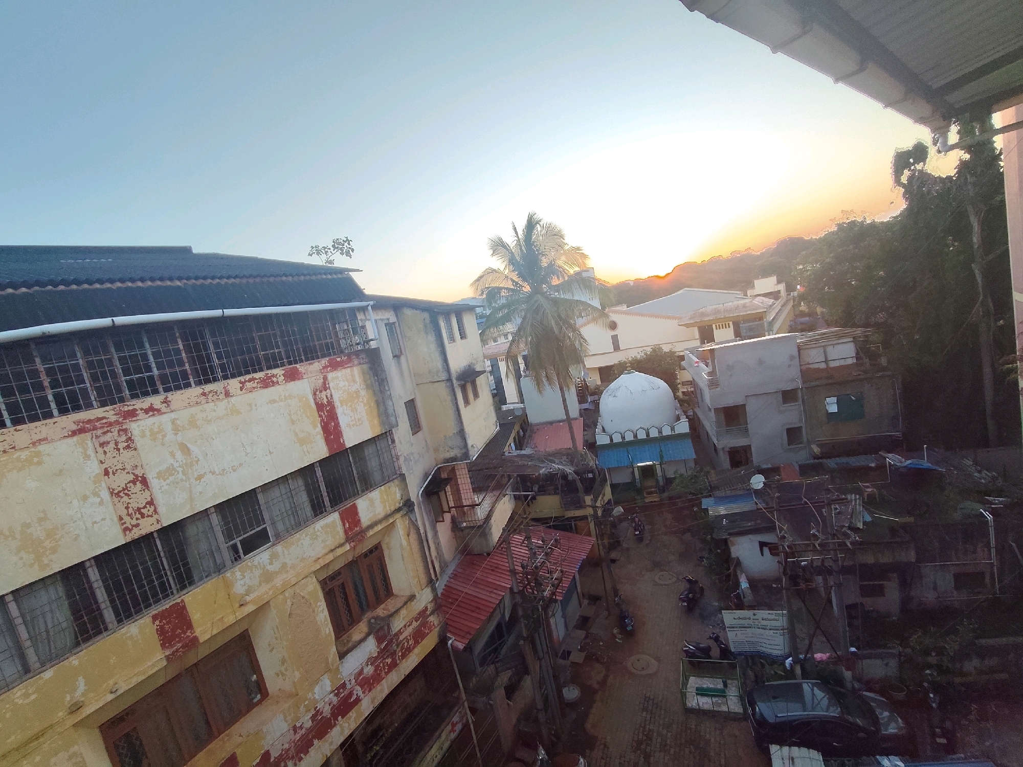 The image captures a view of a narrow street in Belagavi, India, at sunset. The sky is a pale blue, transitioning to orange and yellow near the horizon, with the sun partially obscured by a distant, tree-covered hill. A prominent building on the left, painted cream and red, has numerous windows with metal grills. Across the street, a white-domed structure, likely a mosque, with blue trim and an arched entrance, is visible. Several other buildings of varying heights and architectural styles are present, some with corrugated metal roofs. The street is paved with bricks and appears wet, suggesting recent rain. Parked motorcycles and a dark-colored car are scattered along the street, indicating a residential or mixed-use area. Electrical poles and wires crisscross the scene. The overall impression is of a densely populated urban environment bathed in the warm light of the setting sun.