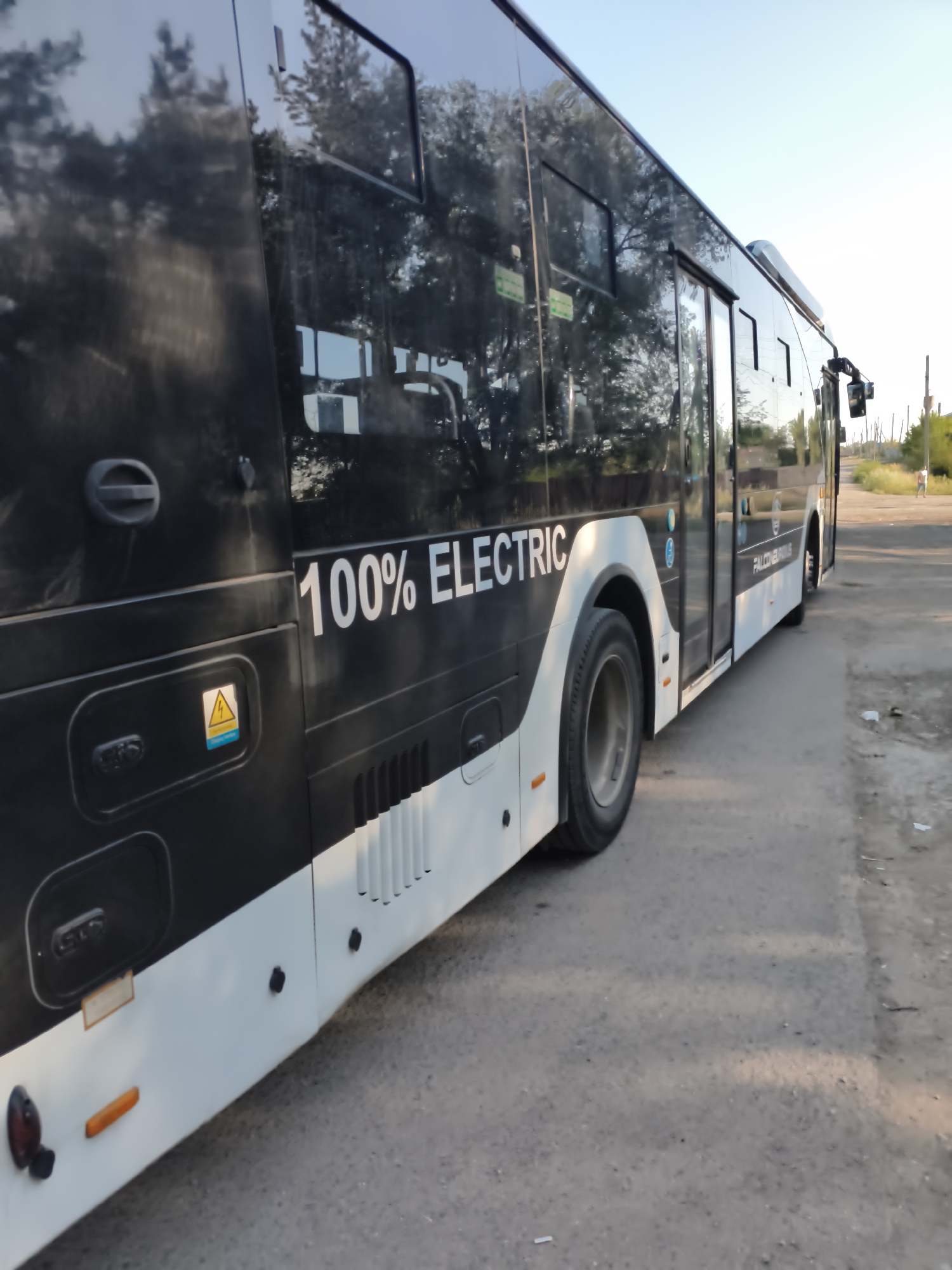 An outdoor, eye-level shot captures the side of a black and white bus parked on an asphalt road. The bus is predominantly black with a white lower body. White text on the bus reads "100% ELECTRIC." A yellow triangular warning symbol with a lightning bolt is visible on the black portion of the bus, accompanied by blue text below it. Several windows line the side of the bus, reflecting trees and the sky. The environment appears to be a roadside with dry grass and sparse vegetation. In the background, a distant figure walks away from the bus on the right side of the frame. The sky is clear and bright.