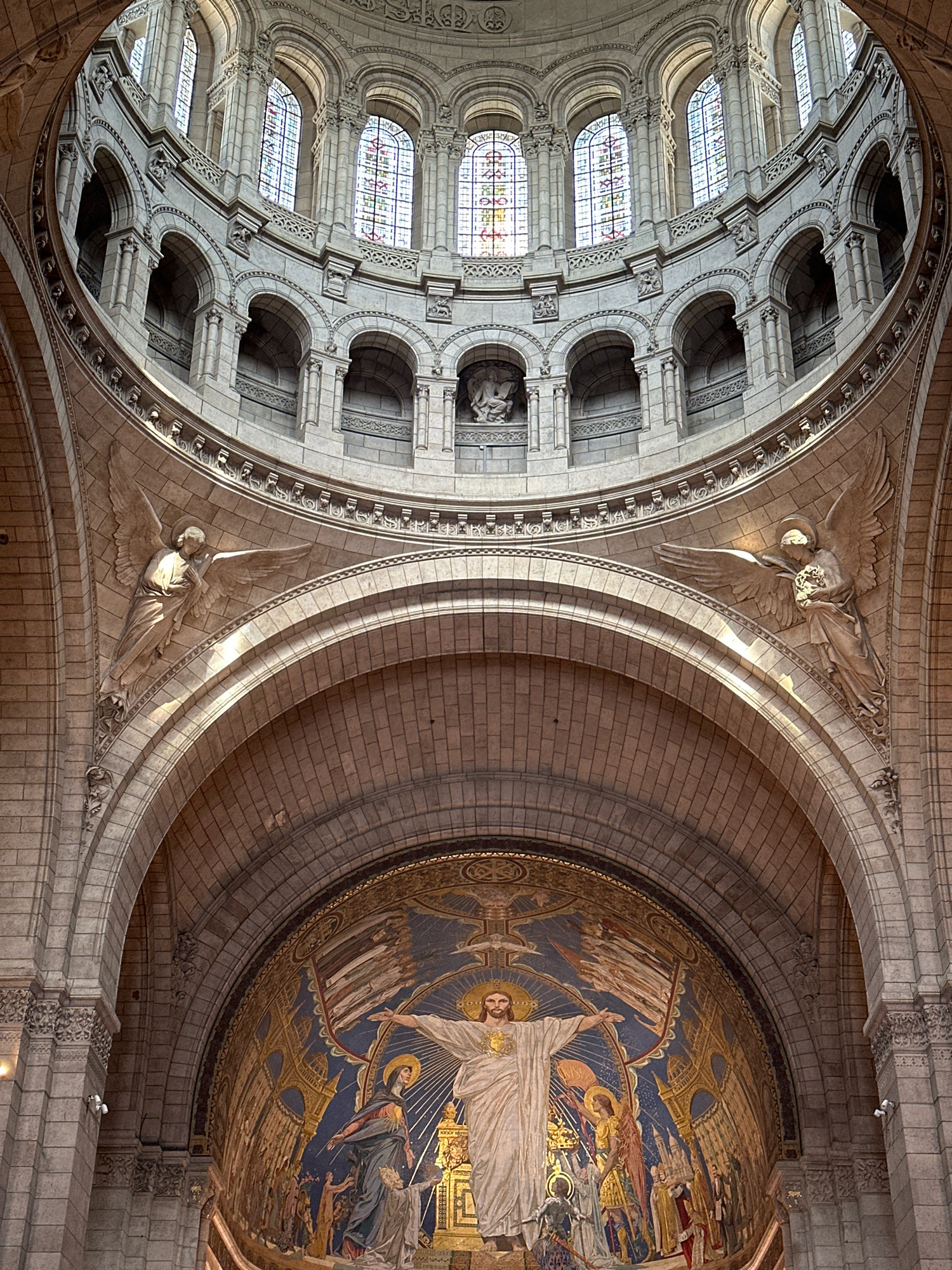 The image shows the interior of the Sacré-Cœur Basilica in Paris, France. The viewpoint is looking upwards towards the dome and apse. Dominating the lower portion of the image is a large, ornate mosaic in the apse depicting a radiant figure of Jesus Christ with outstretched arms, surrounded by other figures, including what appears to be Mary and an angel. The mosaic is set against a vibrant blue background with golden architectural elements and a starry sky. Above the apse, a grand stone archway leads the eye towards the intricate stonework of the dome, which features multiple levels of arched windows with stained glass, supported by columns and decorative carvings. Two large winged angel sculptures are prominently placed on the lower curved walls, facing inwards towards the center. The overall impression is one of grand religious architecture, with a focus on stone masonry, mosaics, and sculpted figures bathed in natural light filtering through the stained-glass windows.