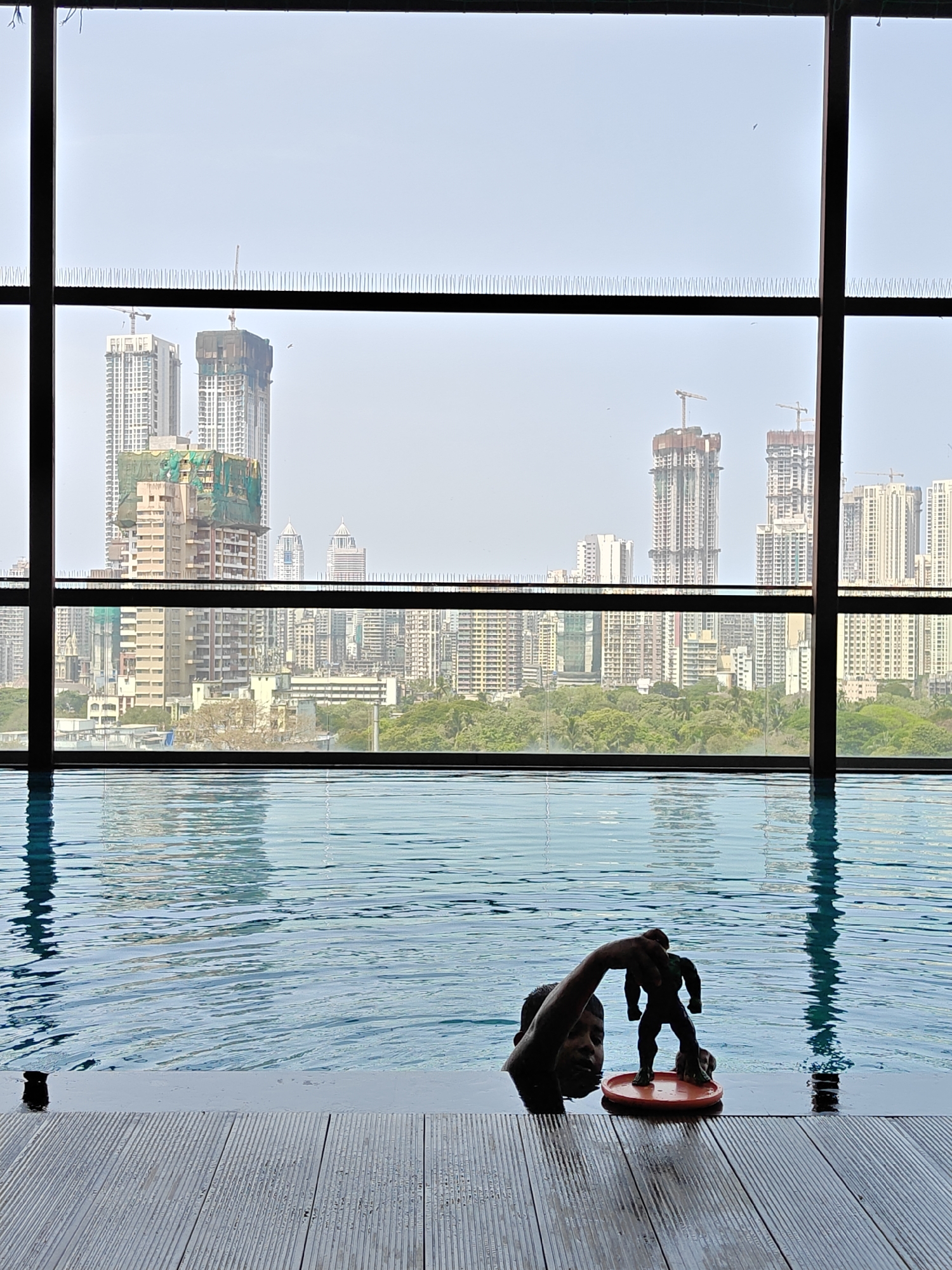 A medium shot reveals a child in a swimming pool, holding an action figure. In the background, through the expansive windows of the pool area, lies a cityscape characteristic of Mumbai, India. The architecture includes various mid and high-rise buildings, some under construction, against a hazy skyline.
The pool's edge is a grey wood-like composite, while the water is rippled and reflects the cityscape above. Anti-bird spikes are visible atop the window frames. The child's upper body is visible in the water, with one arm outstretched, supporting a plastic toy.