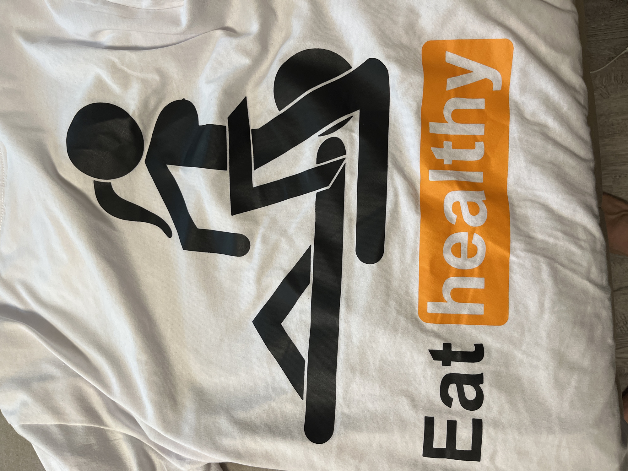 A white, short-sleeved t-shirt, located in Plovdiv, Bulgaria, lies flat on a light brown, textured surface, possibly wood. The shirt features a large, black graphic depicting two stylized human figures in a prone, intimate posture. The upper figure, identifiable as female by a ponytail, is positioned above and behind the lower figure, which is outstretched horizontally on a base. Below the graphic, the text "Eat healthy" is printed. The word "Eat" is in black capital letters. The word "healthy" is rendered in white capital letters within a rectangular orange box with rounded corners. The fabric of the t-shirt exhibits minor creases and folds. A small portion of a human foot, with visible toes, is partially visible at the bottom left edge of the frame. The scene is illuminated by what appears to be ambient indoor lighting.